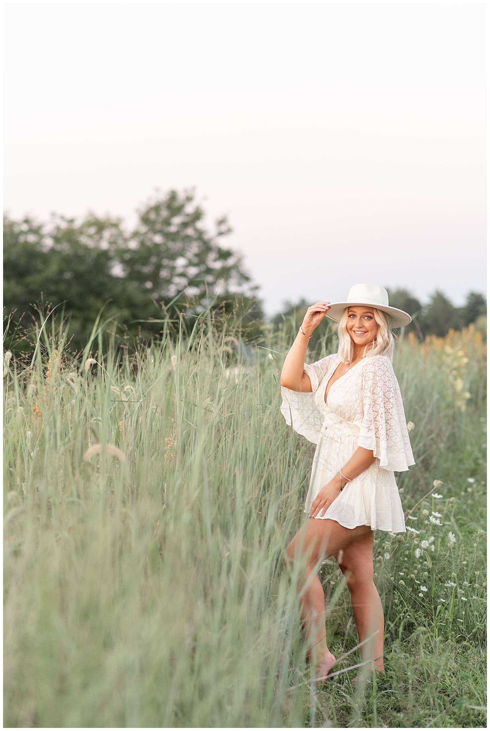 senior girl standing at edge of tall wild grasses with her right hand holding brim of white hat with left shoulder toward camera at overlook park at sunset