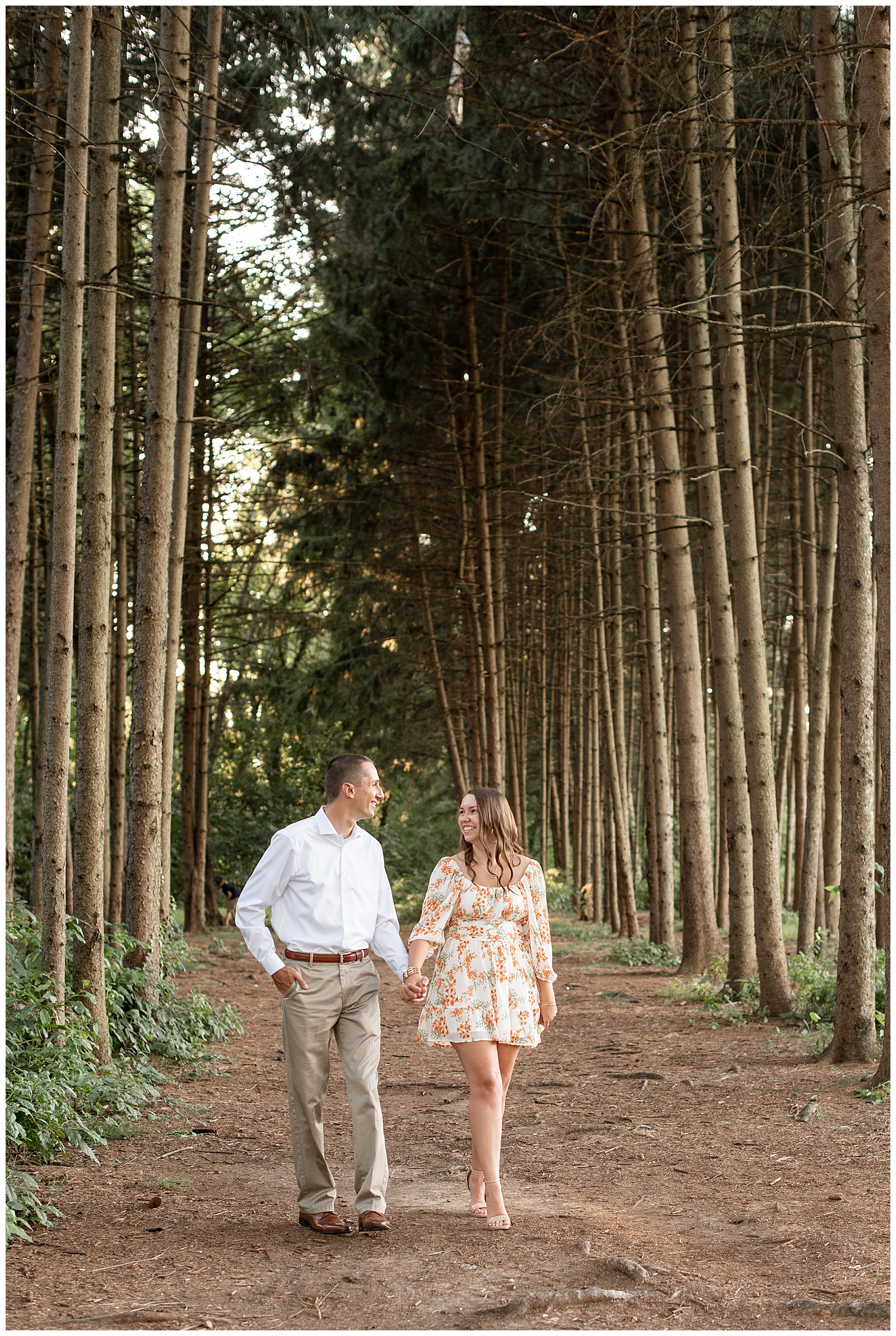 guy wearing white button up shirt and khaki pants and girl wearing floral dress holding hands between rows of evergreen trees at overlook park