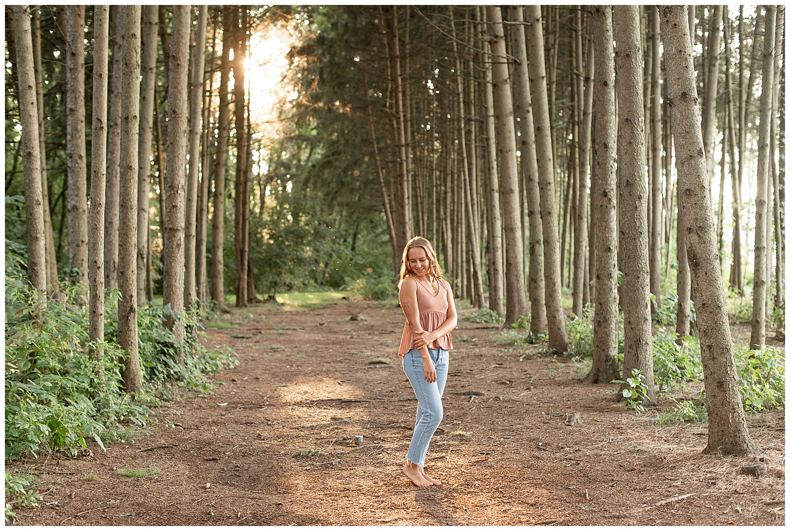 senior girl wearing peach tank top and blue jeans standing between evergreen tree rows at overlook park
