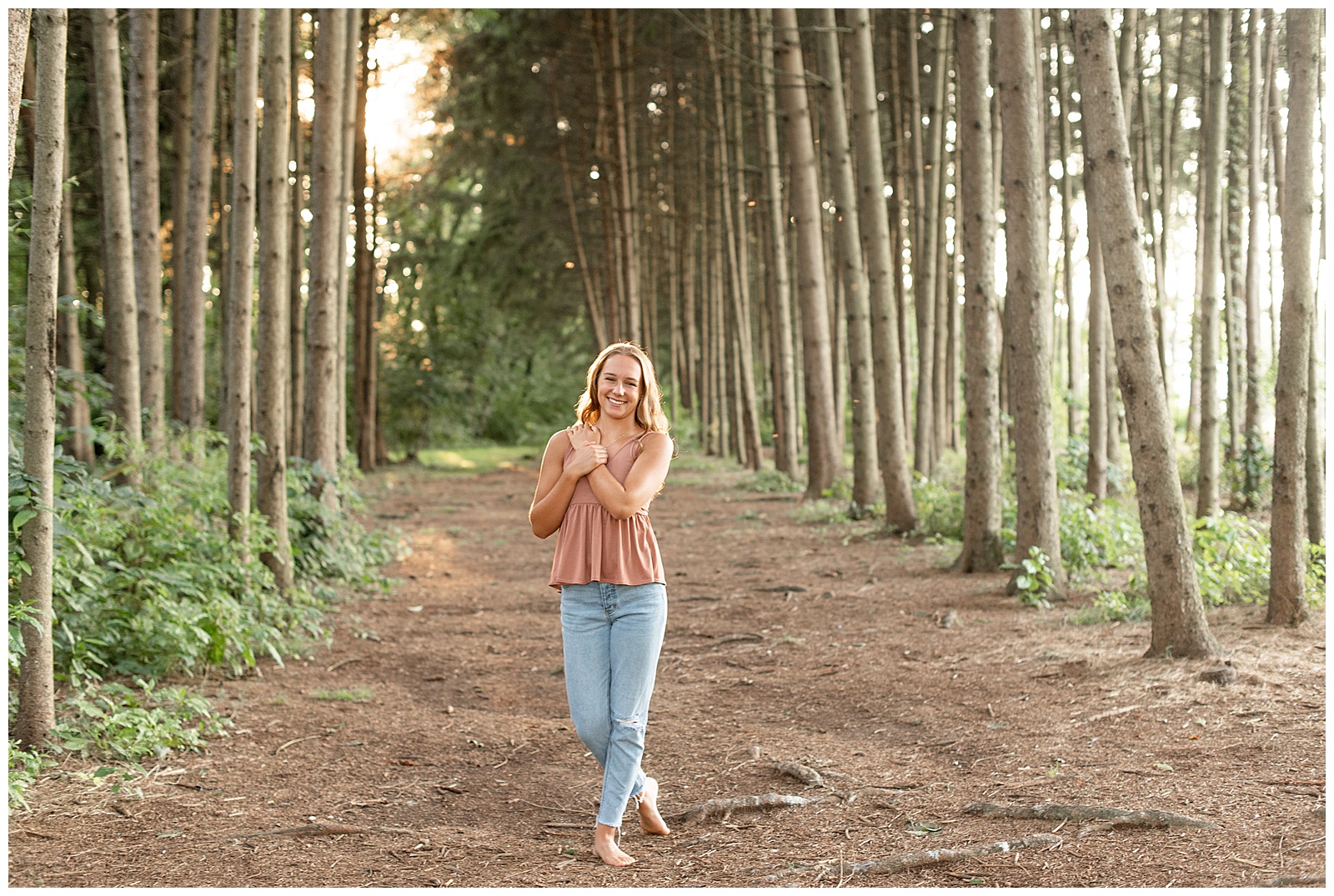senior girl with left hand on right shoulder and right toe back by evergreen trees at park in lancaster county