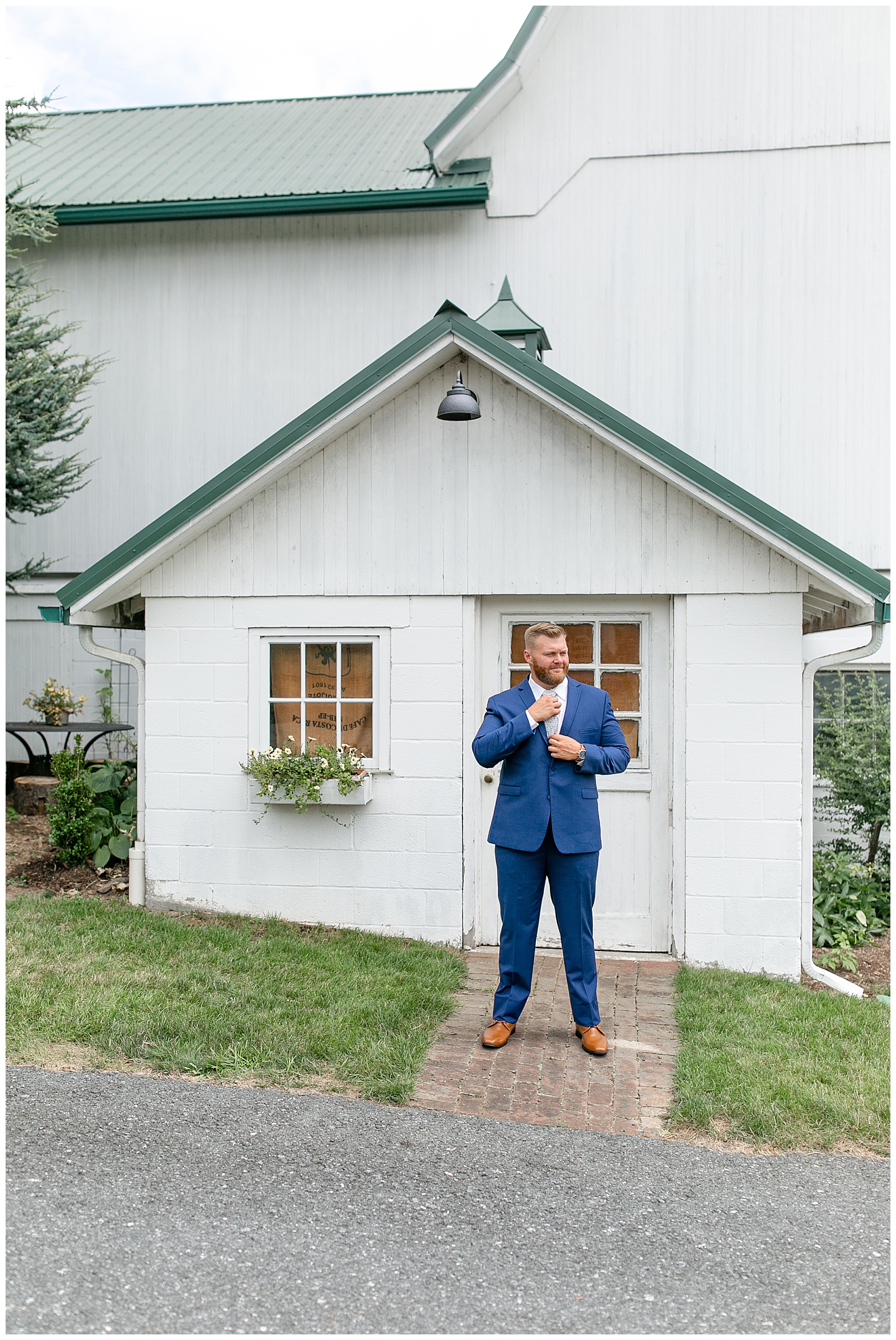 groom adjusting his tie as he stands by white barn wearing blue suit at lakefield weddings