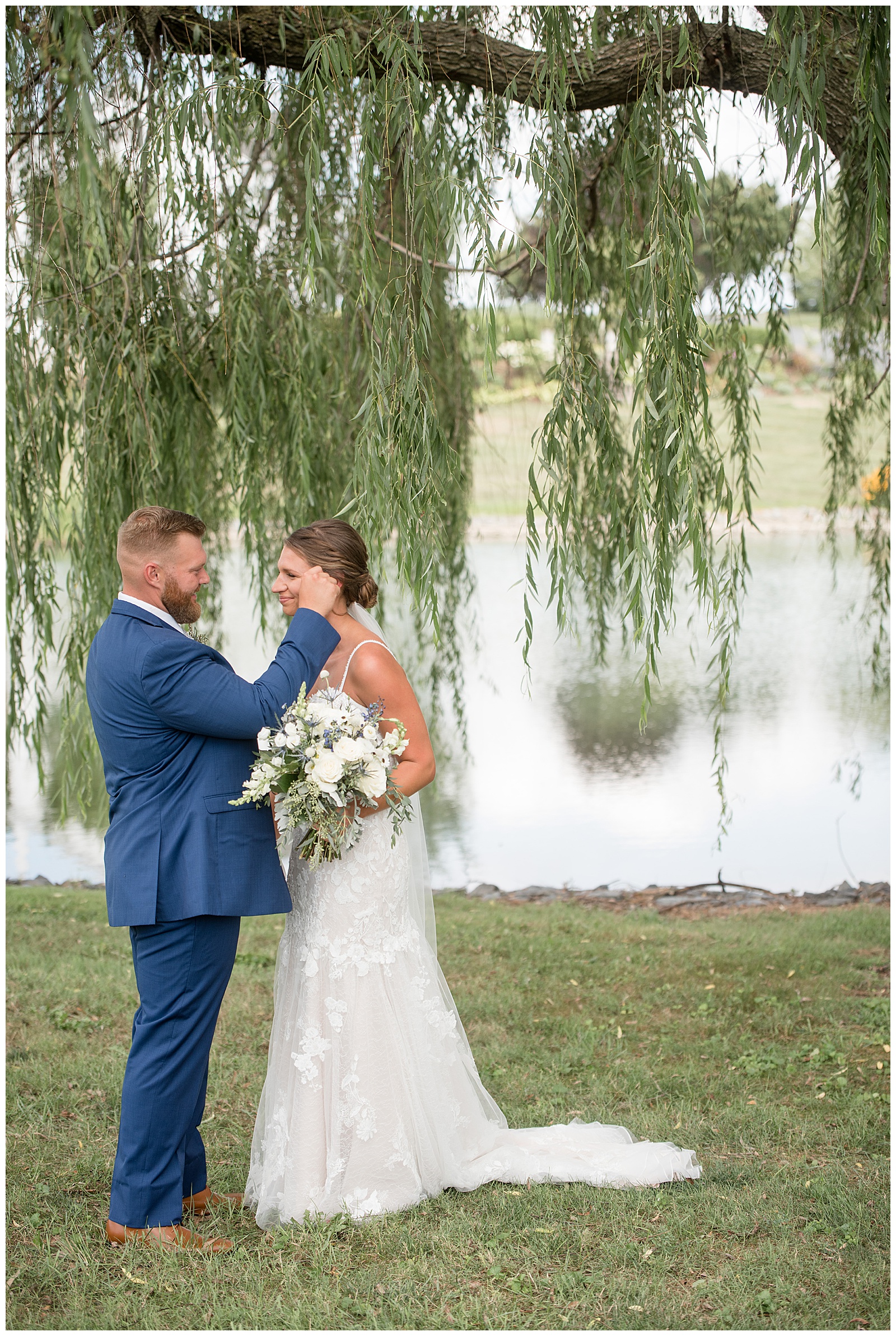 groom wiping bride's tear during first look moment by lake and willow tree at lakefield weddings