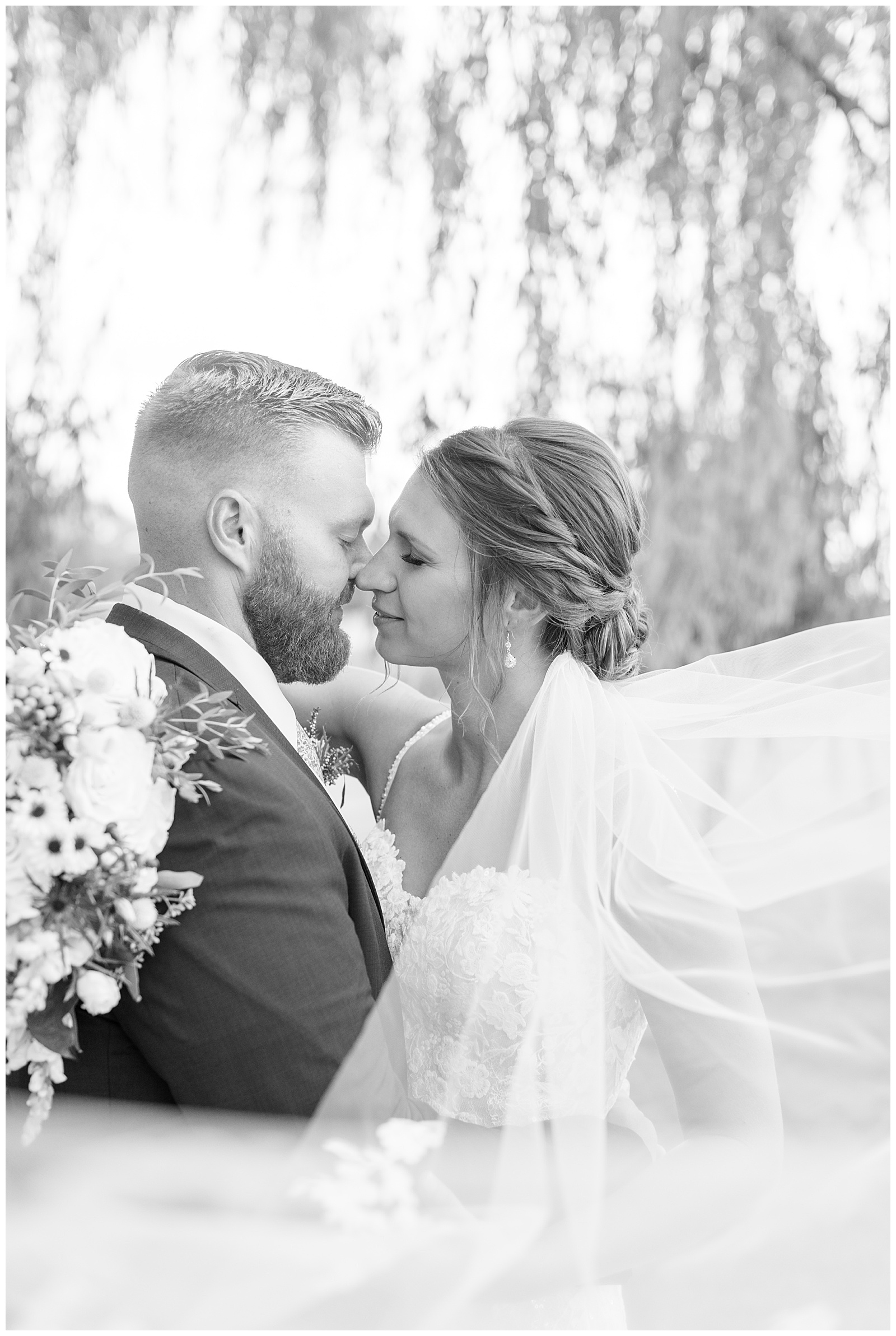 black and white photo of bride and groom almost kissing while they hug by willow tree at lakefield weddings