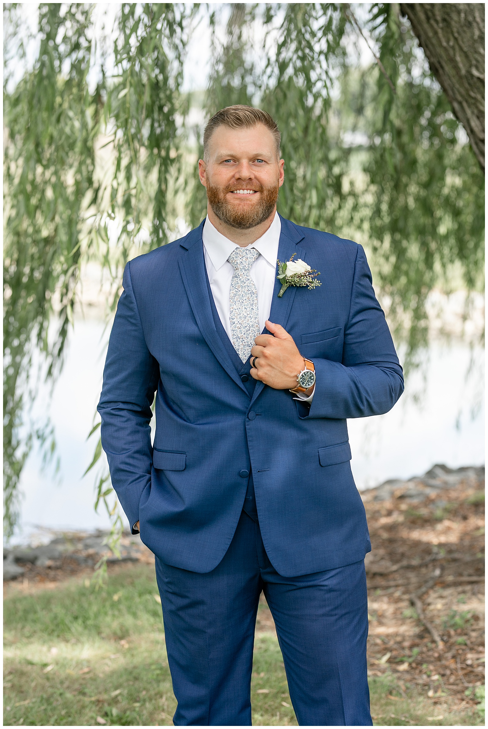 groom smiling as he holds edge of suit coat with his left hand by willow tree at lakefield weddings