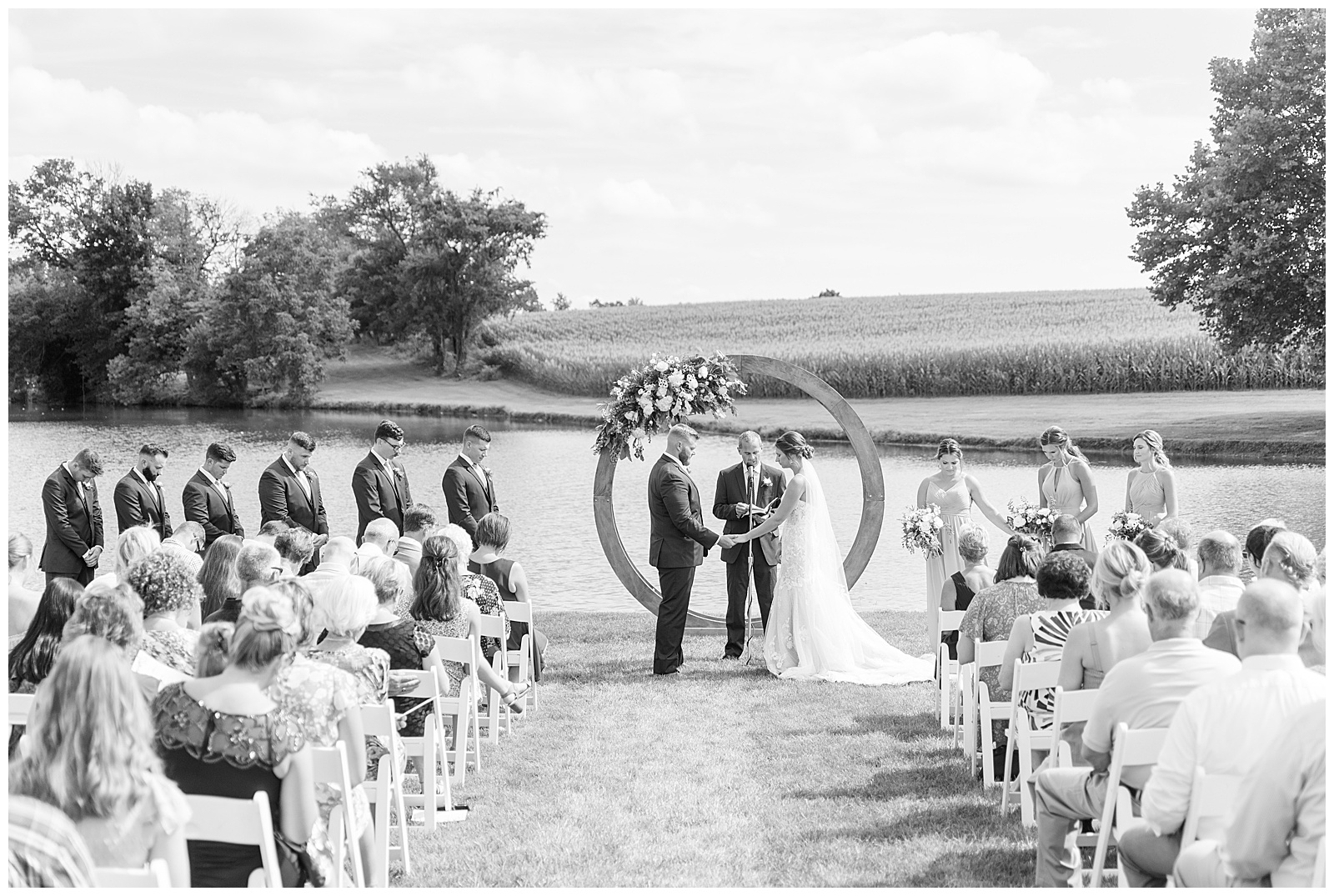 black and white photo of couple sharing their vows by wooden display at pond as guests watch at lakefield weddings