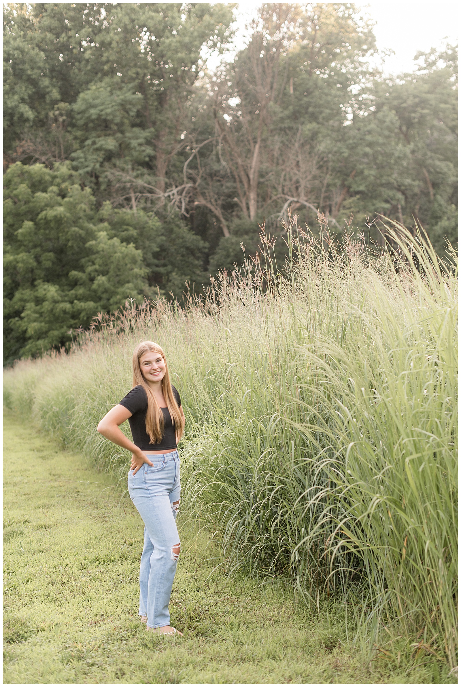 senior girl with hands on hips by tall grasses wearing blue jeans and black t-shirt at hibernia park