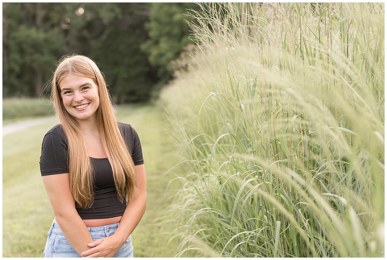 senior girl with left hand holding right arm by tall wild grasses on summer night at hibernia park