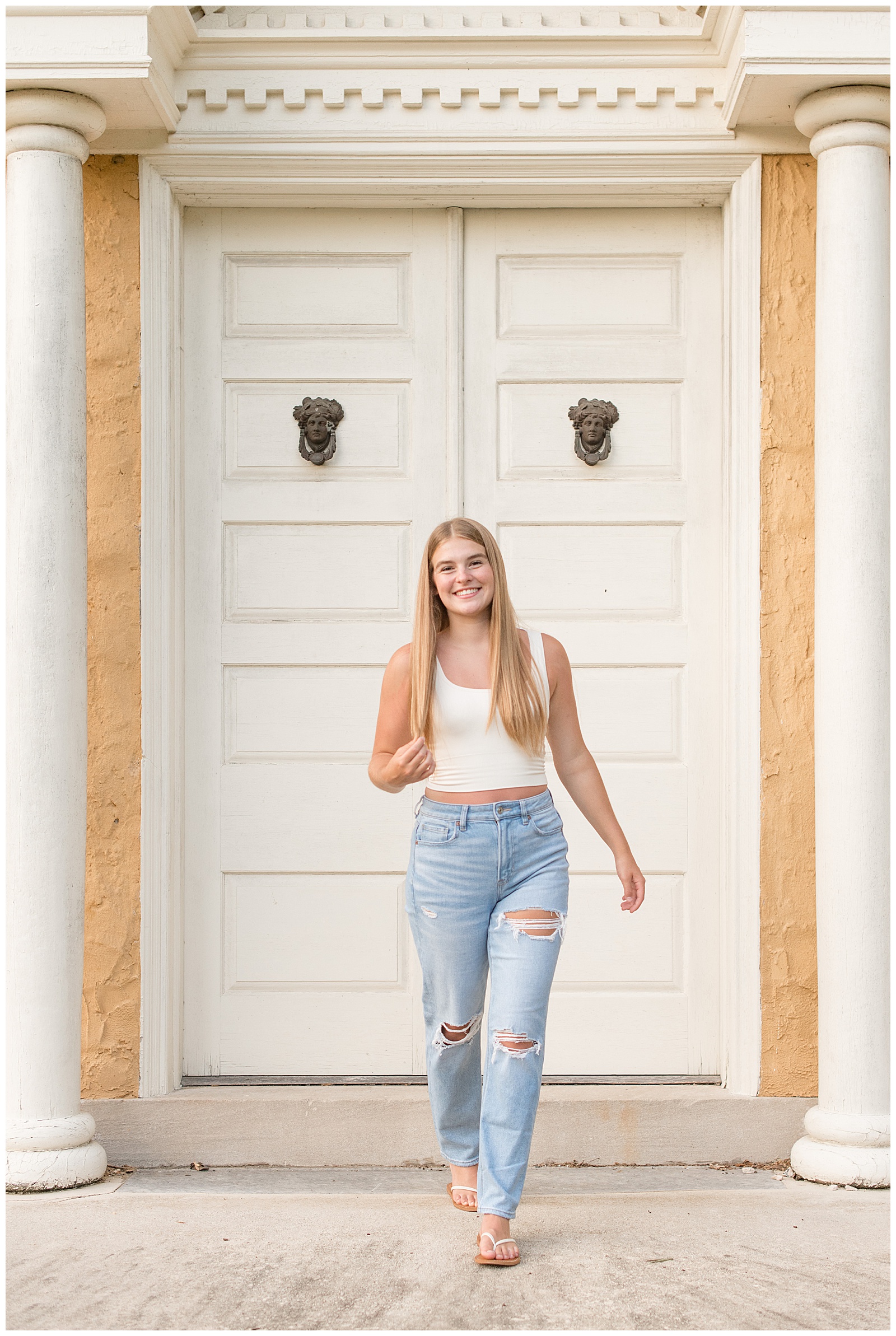 senior girl wearing blue jeans and white tank top walking towards camera and hold tip of her long blonde hair at hibernia park
