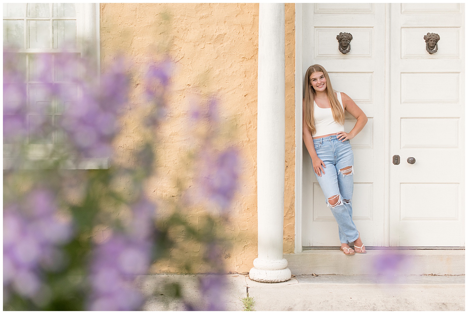 senior girl leaning against white door frame of building with lavender bush in photo at hibernia park