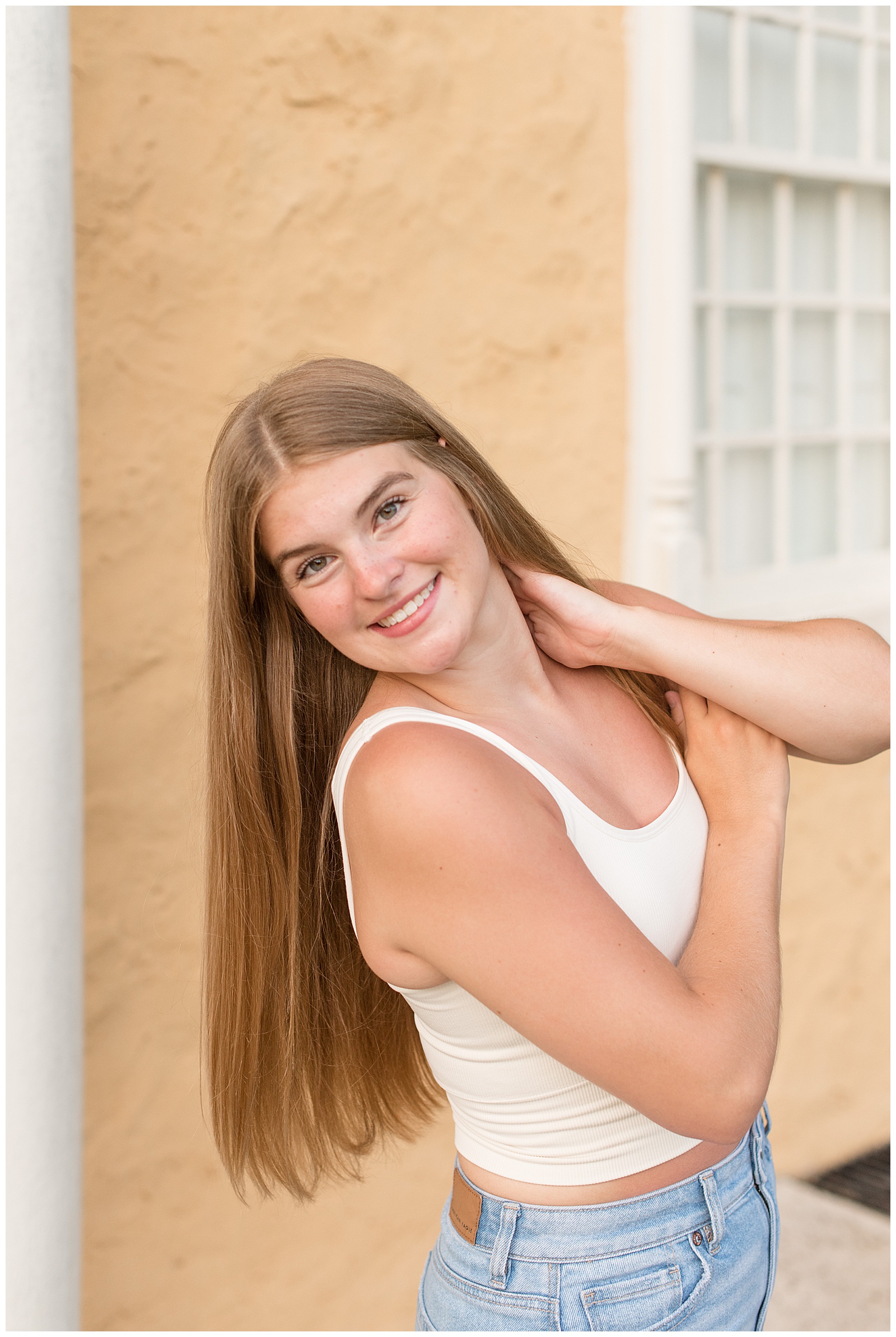 senior girl posing with arms folded and left hand resting on neck by peach colored building at hibernia park
