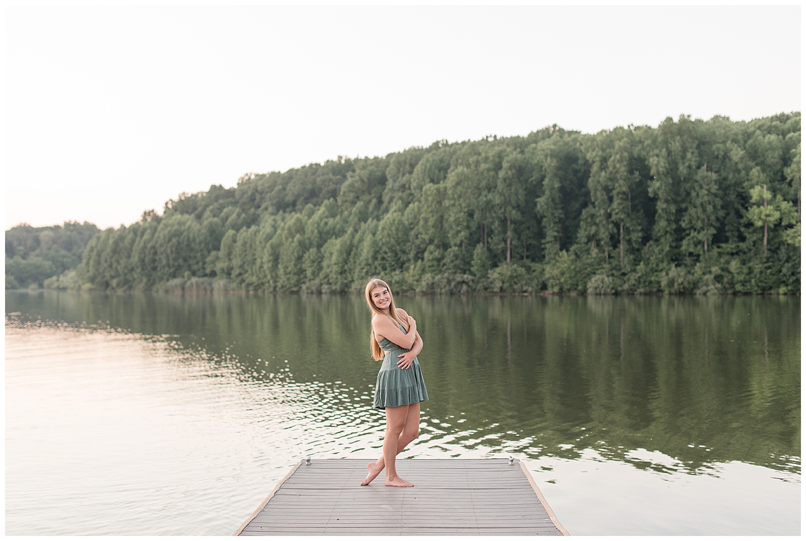 senior girl posing at edge of dock with arms folded over chest and lake behind her at hibernia park