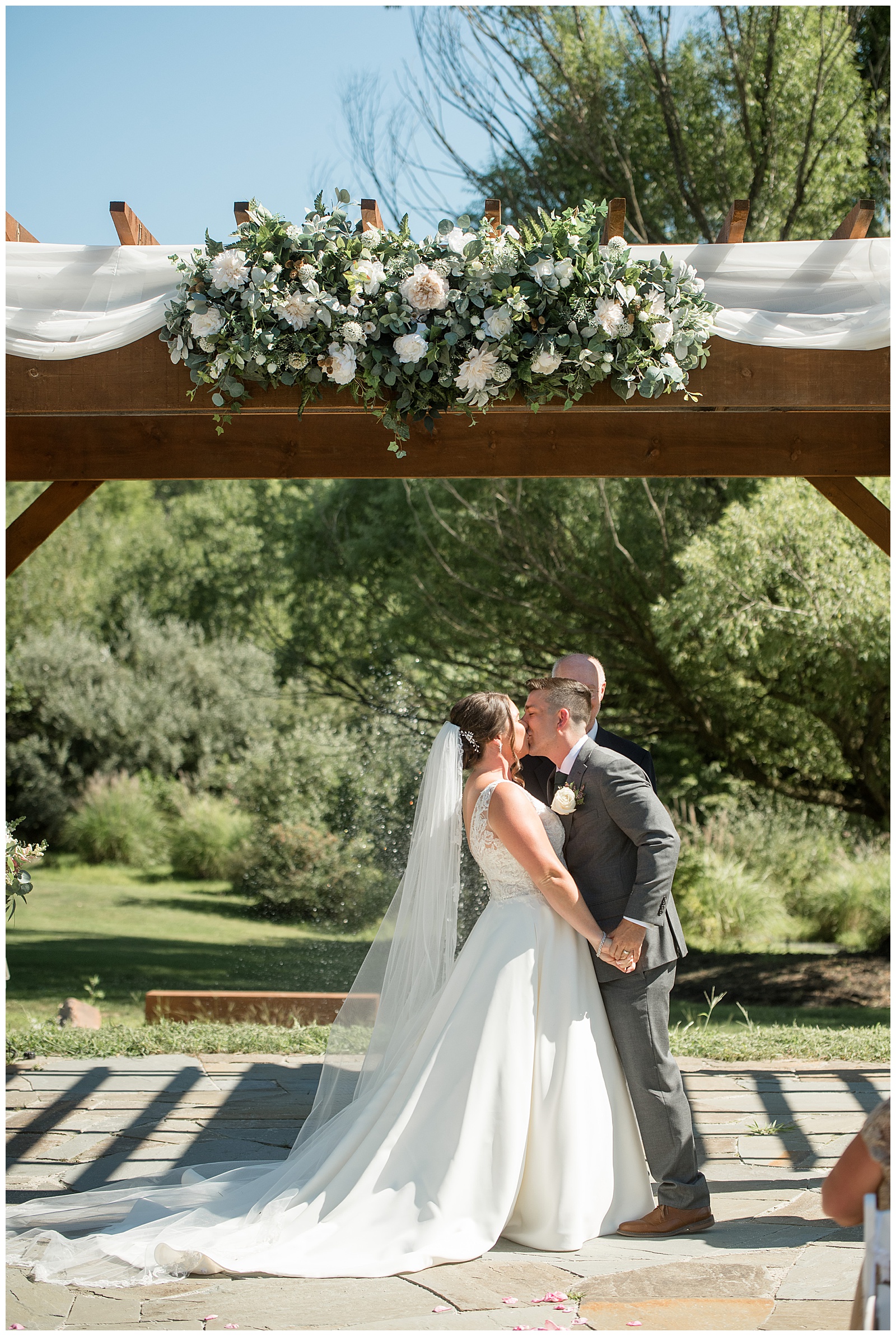 couple sharing their first kiss during outdoor wedding ceremony under wooden display with flowers and white linen at the manor at mountain view