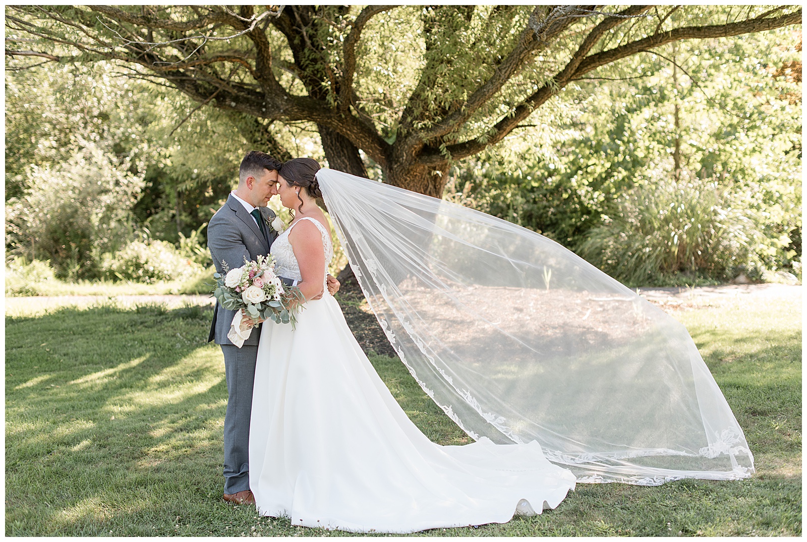 couple hugging with foreheads touching as bride's long white view blows behind her under tree at the manor at mountain view