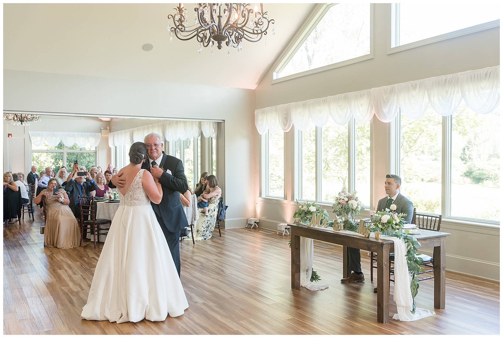 bride dancing with her father as her groom watches them inside white reception room at the manor at mountain view