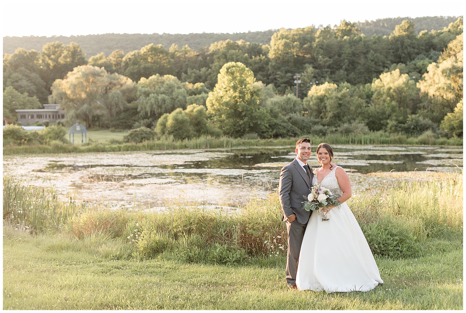 couple standing on right side of photo with large pond and trees behind them at sunset at the manor at mountain view