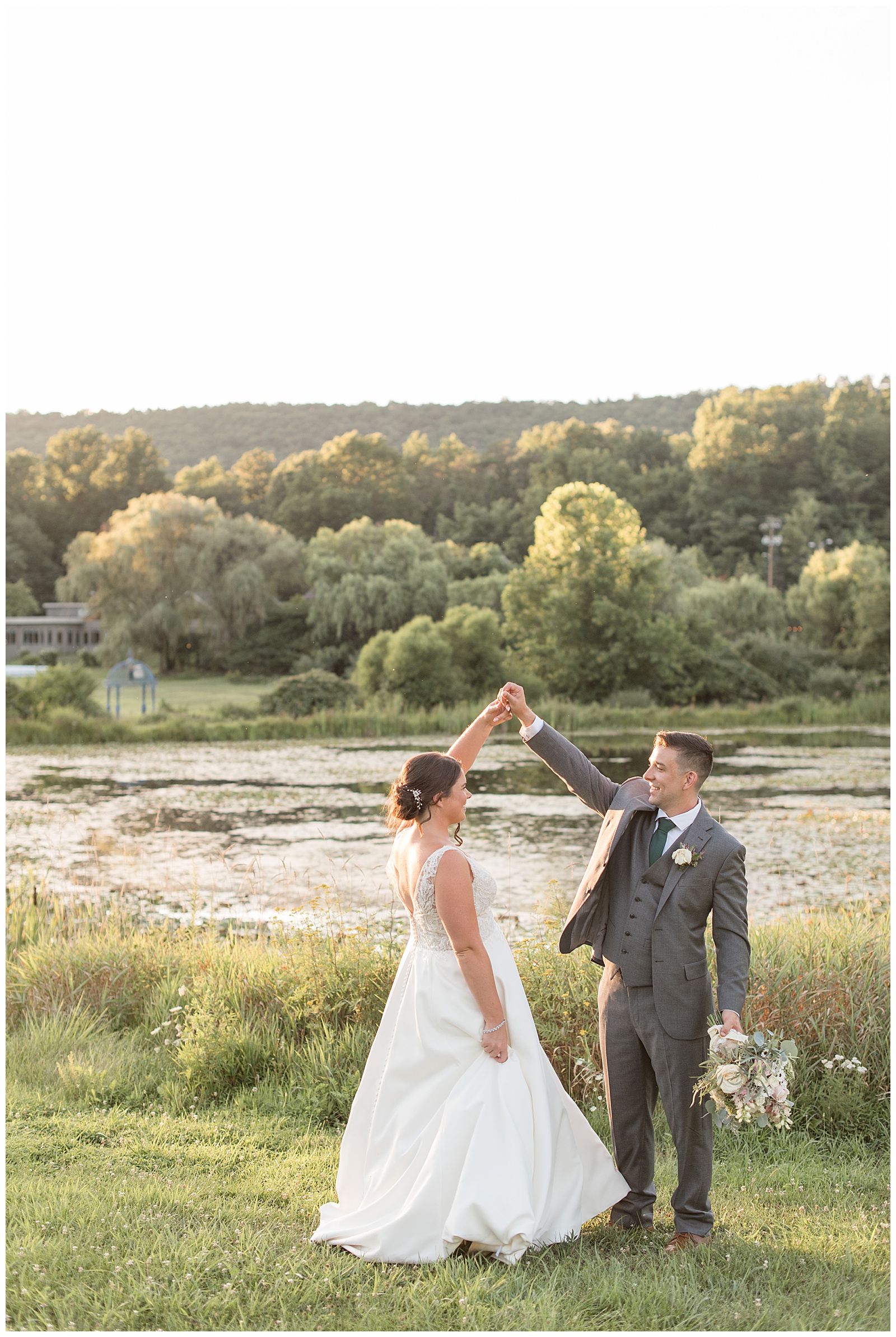 groom twirling his bride under his right hand by pond and trees at sunset in harrisburg pennsylvania