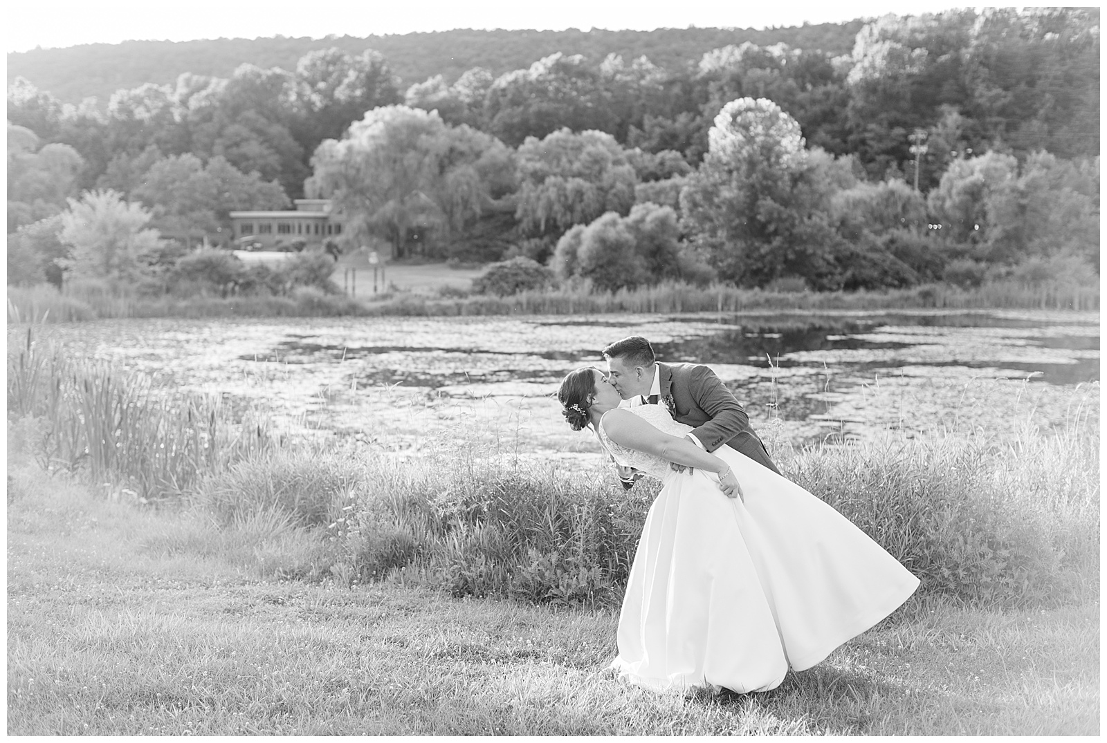 black and white photo of groom dipping his bride back as they kiss by pond at the manor at mountain view