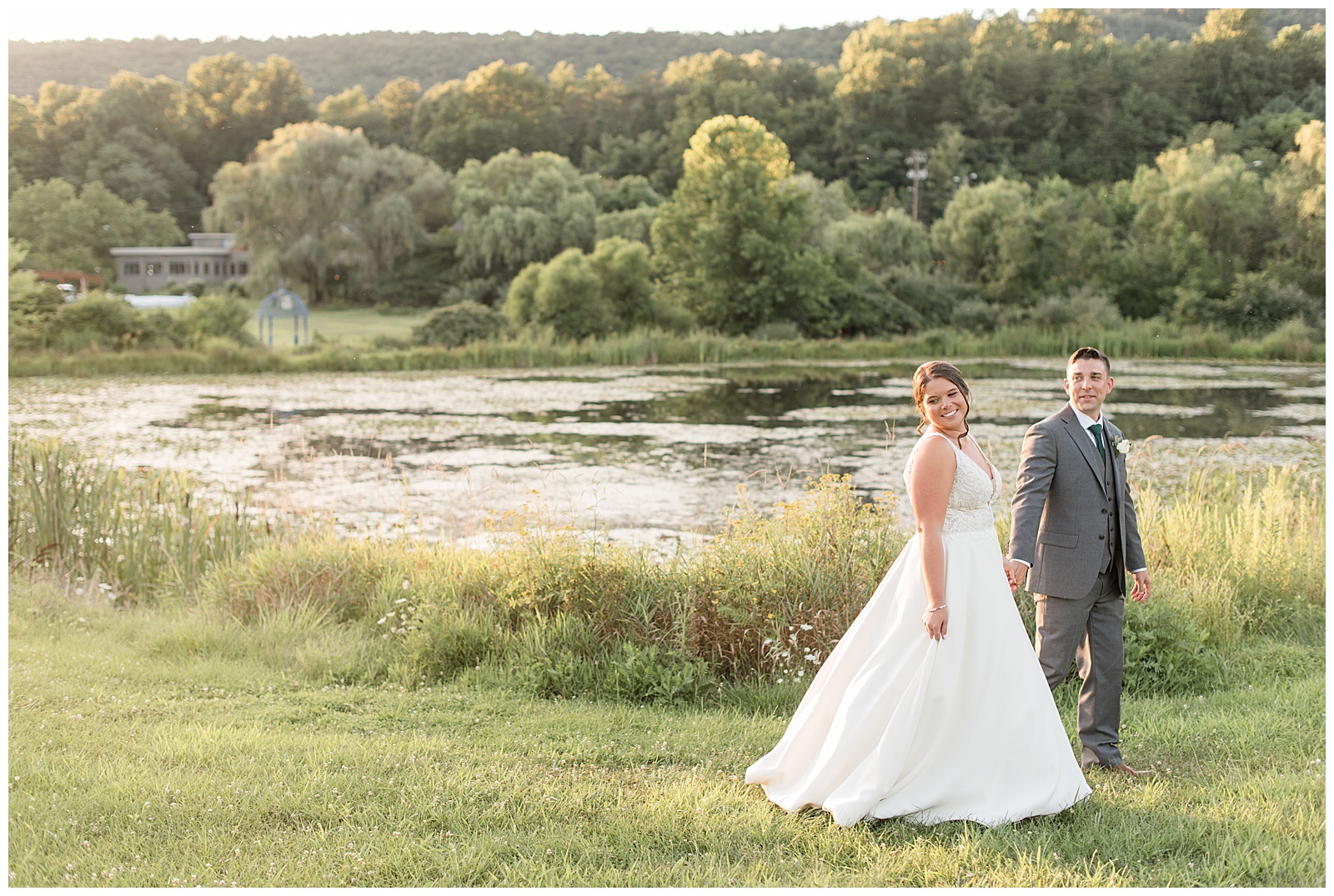 bride and groom holding hands as they walk off camera and look back over right shoulders at sunset by pond at the manor at mountain view