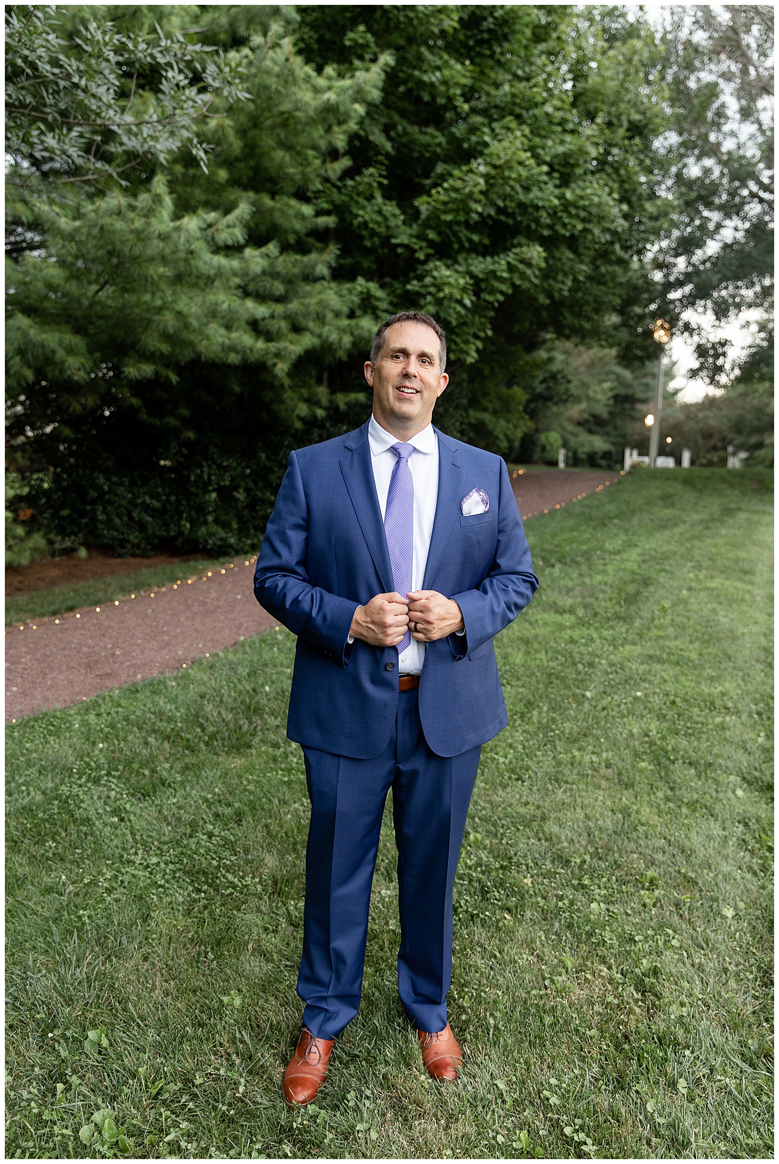 groom in navy blue suit with blue tie holding edges of suit coat in lawn at the inn at leola village