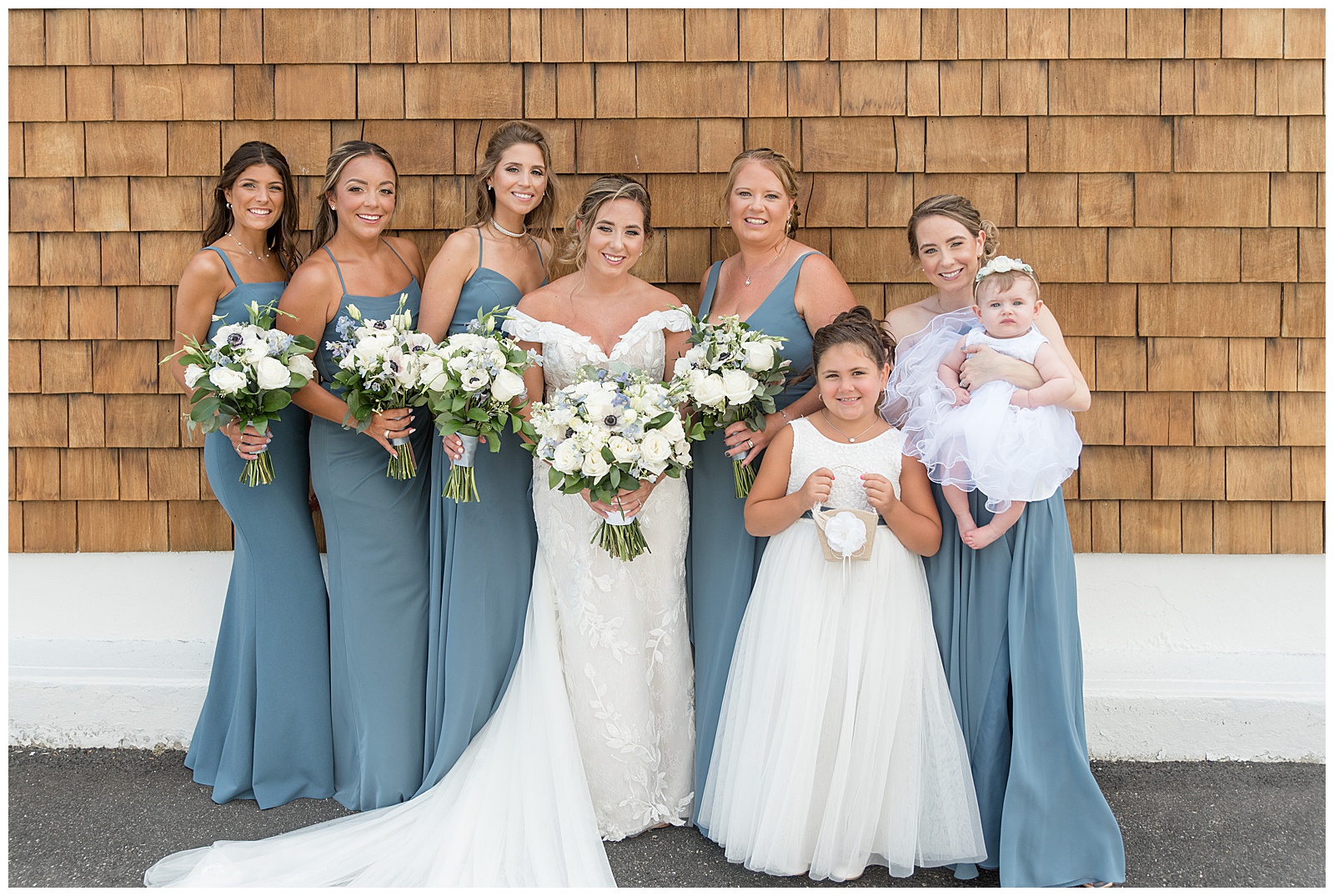 bride in off-the-shoulder white gown surrounded by her bridesmaids and flower girls by tan building in long island, new york