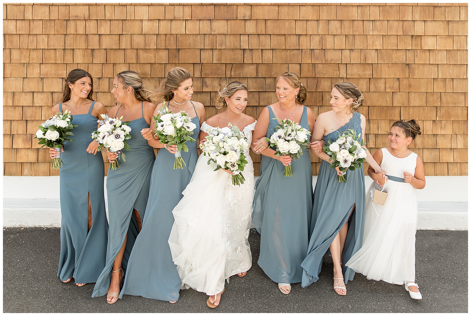 bride arm-in-arm with her bridesmaids walking down driveway by tan building at captain bill's in bay shore, new york