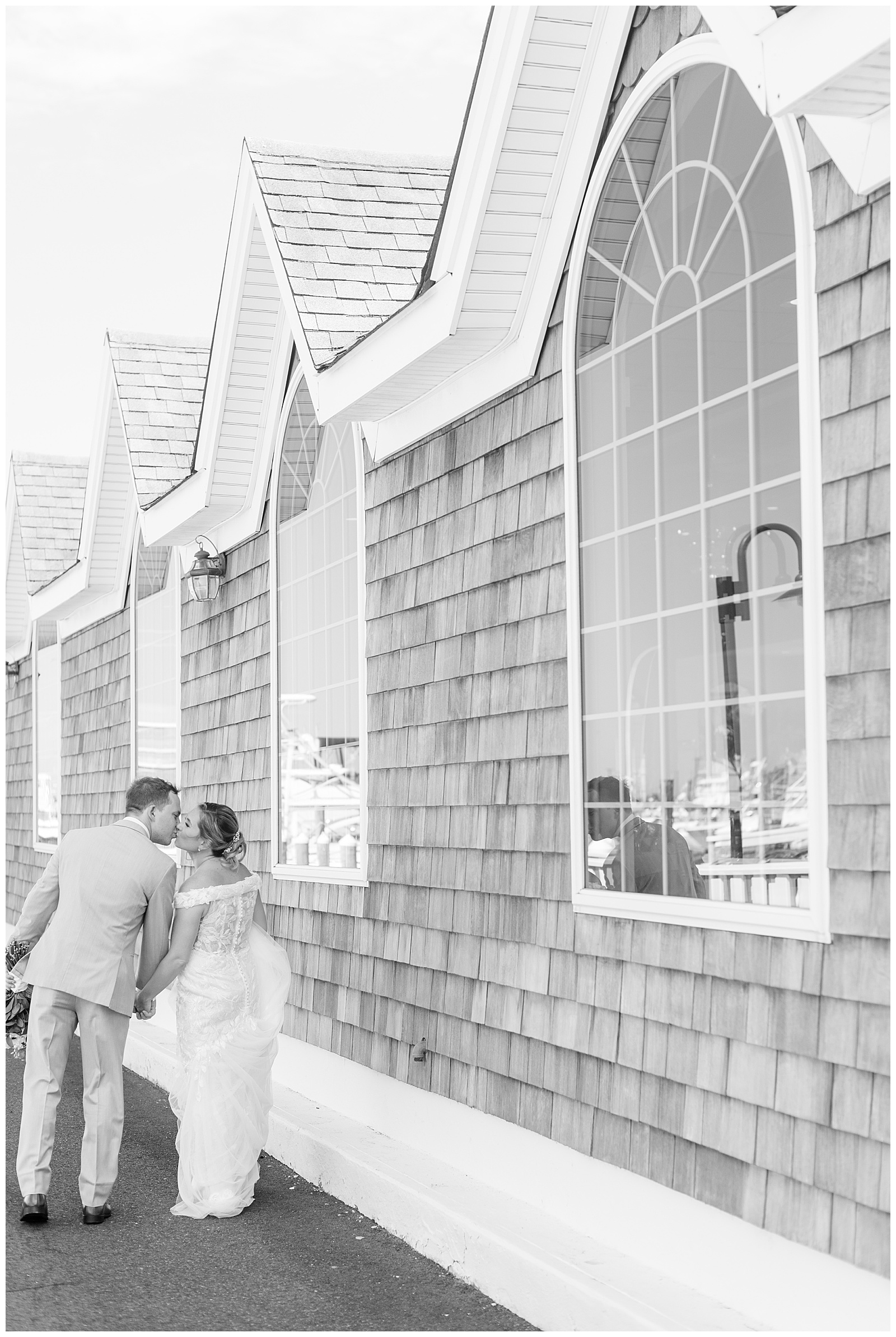 black and white photo of couple kissing as they walk away from camera by waterfront building in new york