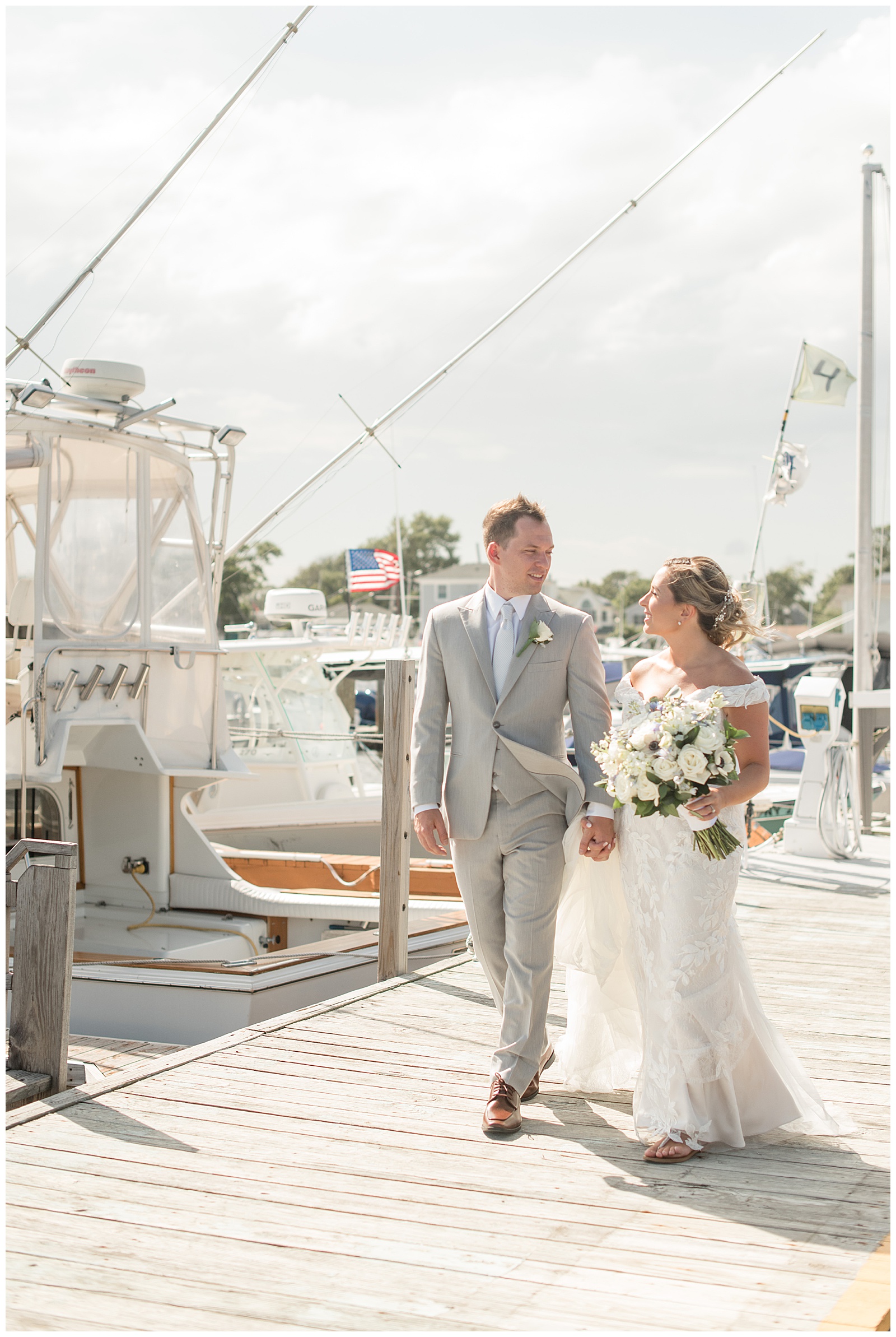 bride and groom holding hands and looking at each other as they walk along dock filled with boats on sunny day in bay shore new york