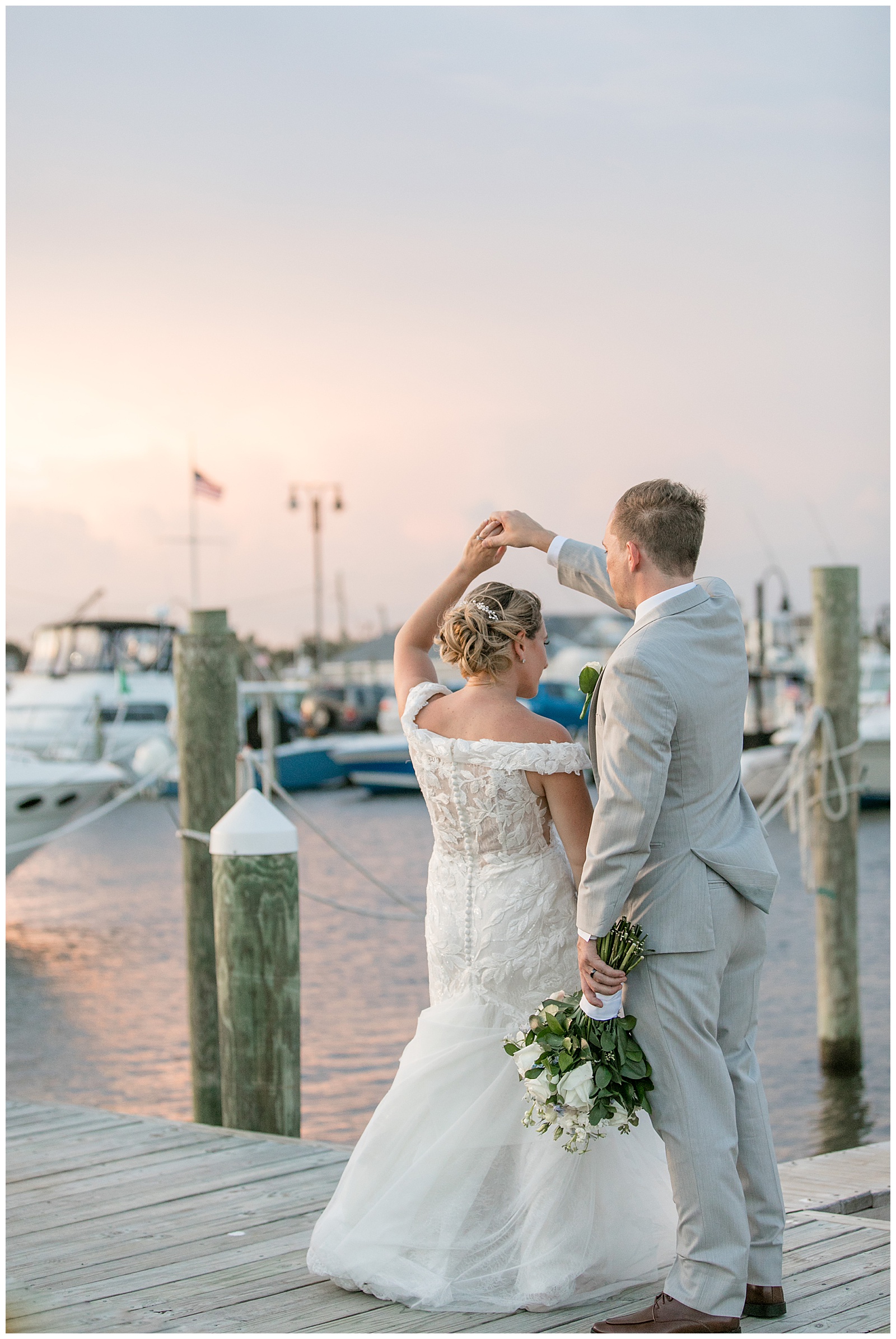 groom twirling his bride under his right hand on dock with boats and the sunset behind them in long island new york