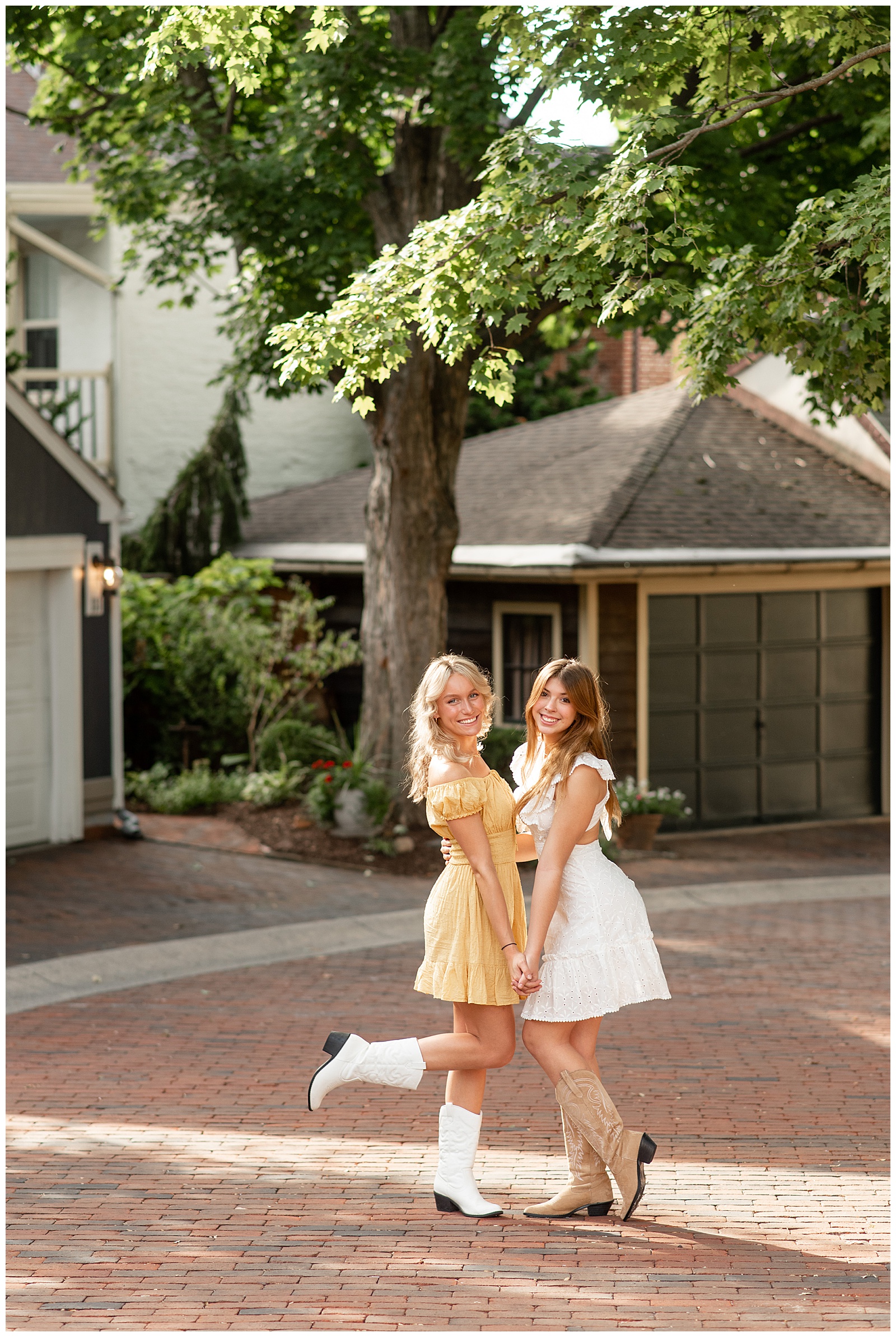 one senior girl in a yellow dress with white cowboy boots and another senior girl in white dress and cowboy boots facing each other holding hands in downtown lancaster