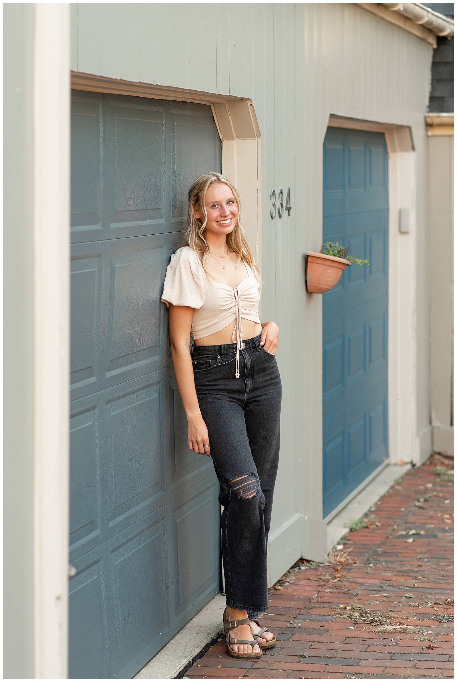 senior girl with left hand in pocket of black jeans smiling at camera and leaning against dark blue garage door in lancaster city