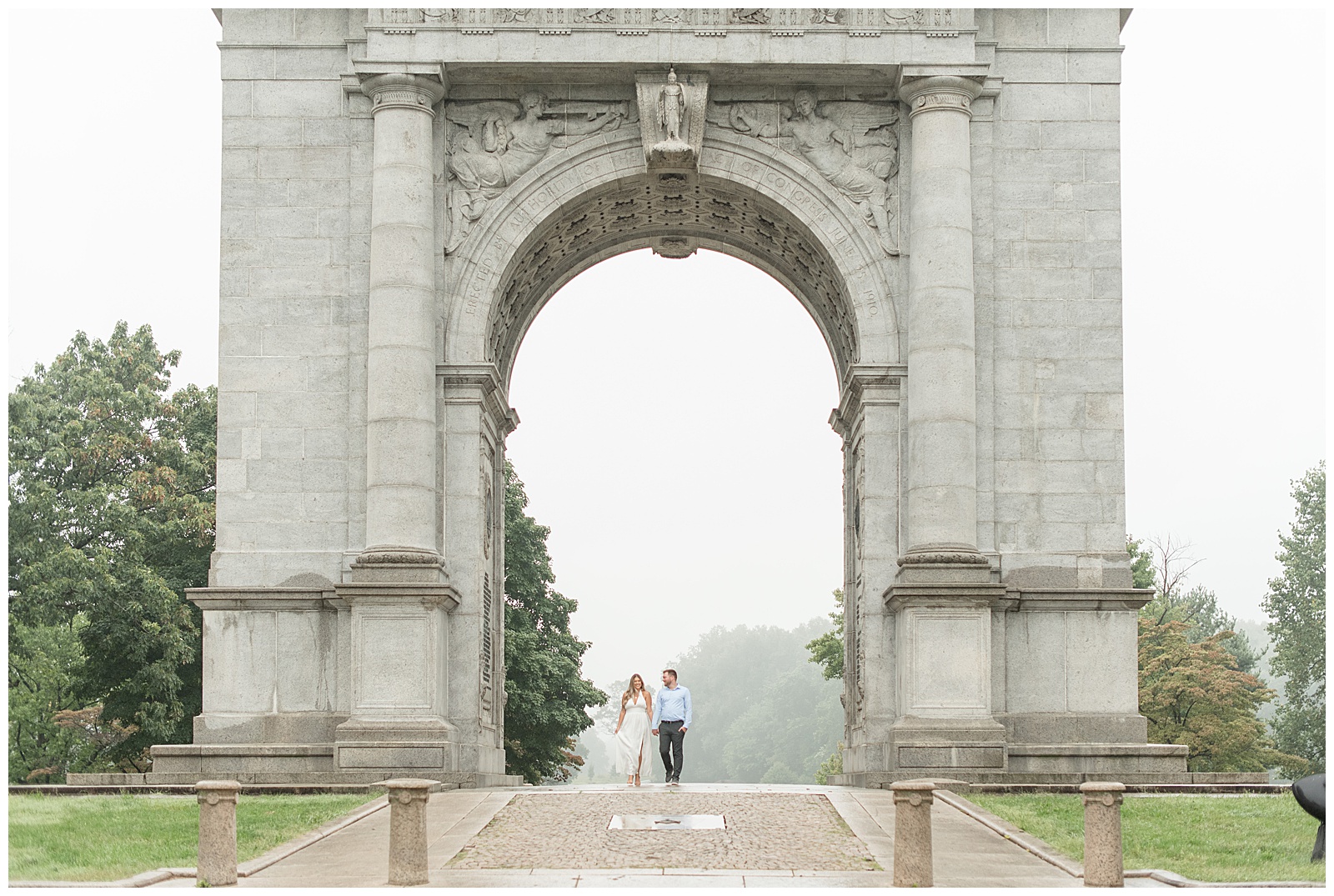engaged couple holding hands and standing underneath national memorial arch on cloudy day in king of prussia