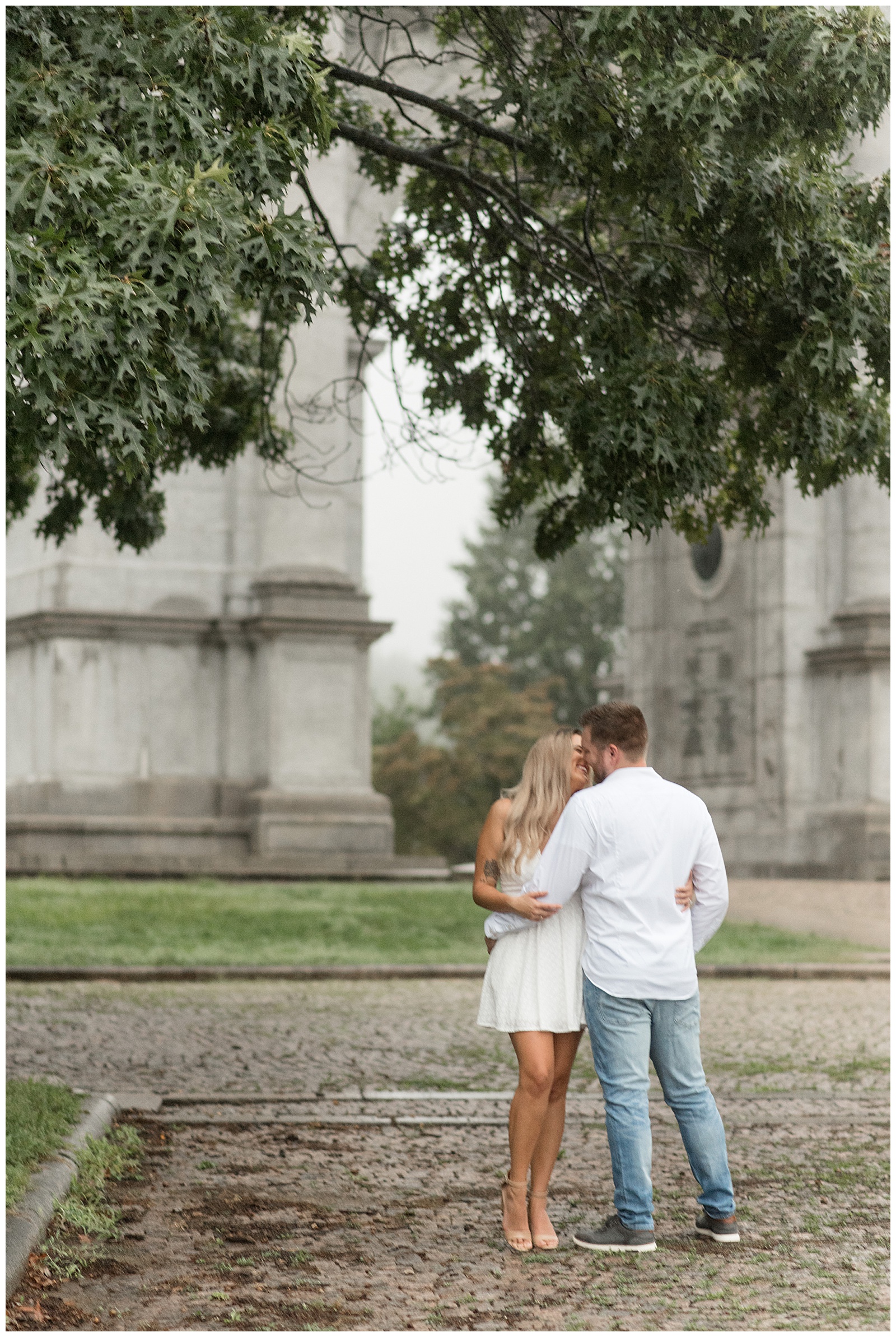 couple hugging with guy's back towards camera as they kiss under a tree with national memorial arch behind them on rainy evening