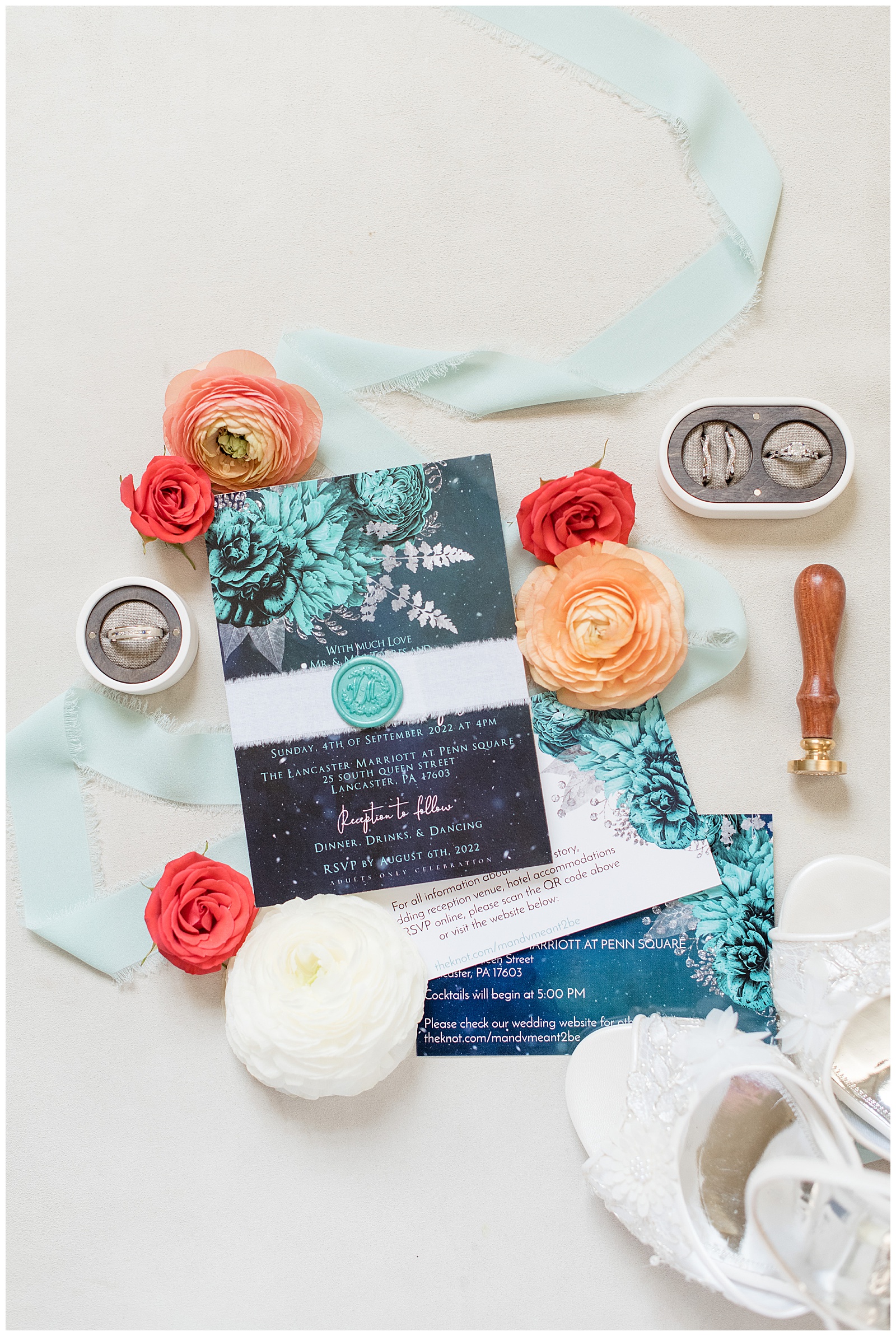 black wedding invitation with white and teal lettering surrounded by colorful ribbon, flowers, and wedding rings in cute display boxes in lancaster pa