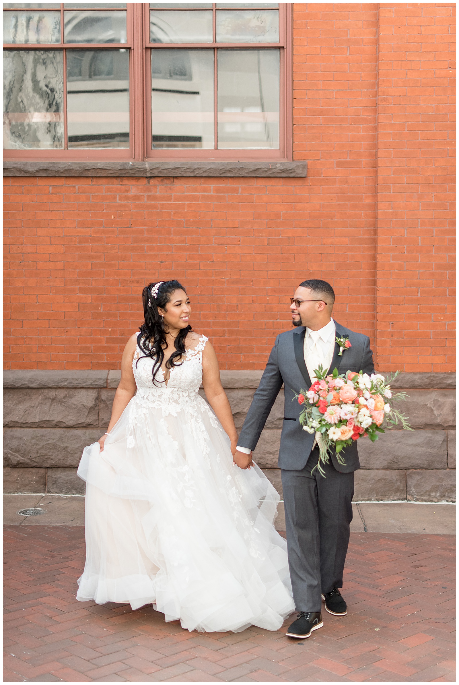 bride and groom holding hands and looking at each other as groom holds colorful bouquet by central market's brick building