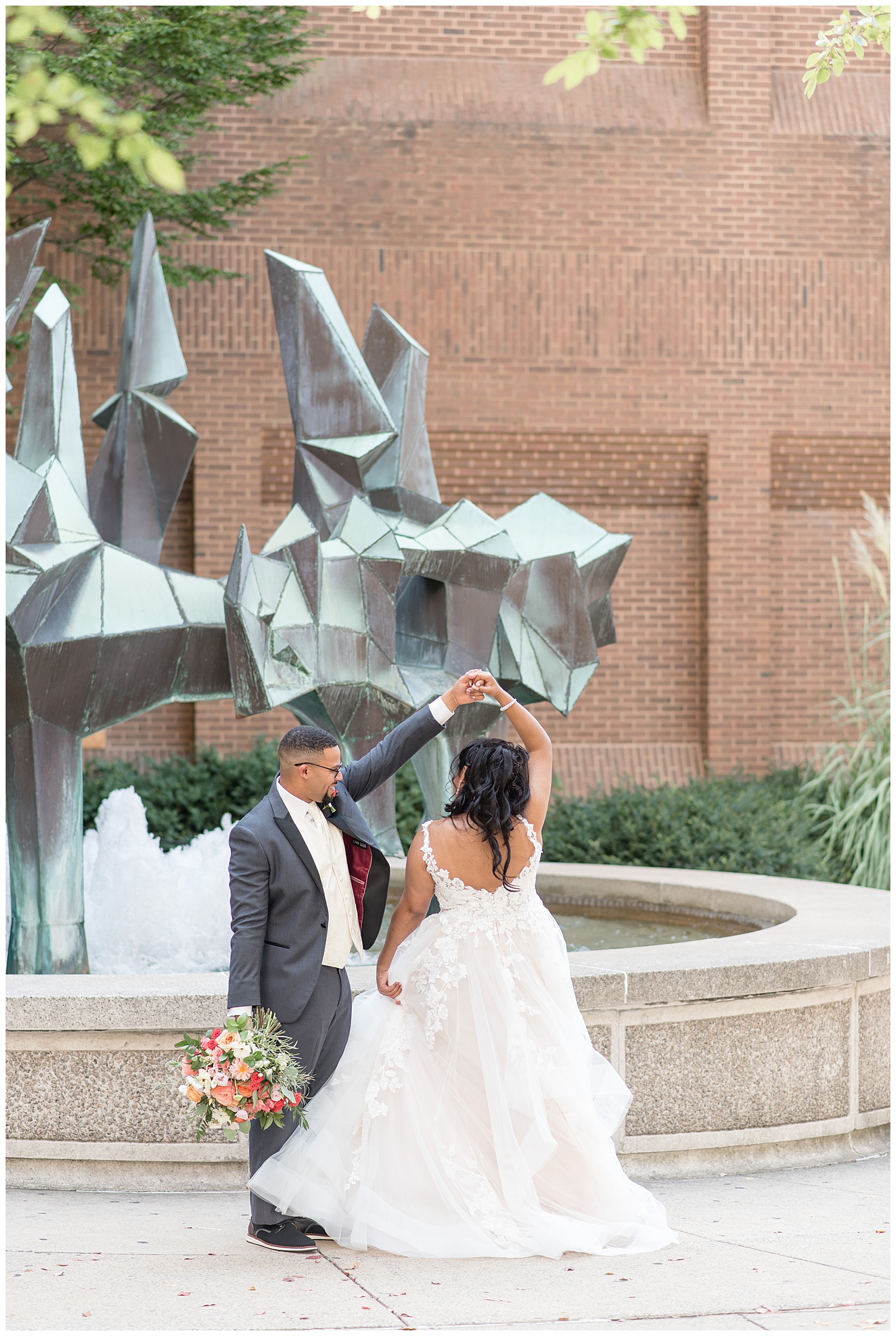 groom holding bouquet and twirling his bride her his left arm by water fountain in downtown lancaster pennsylvania