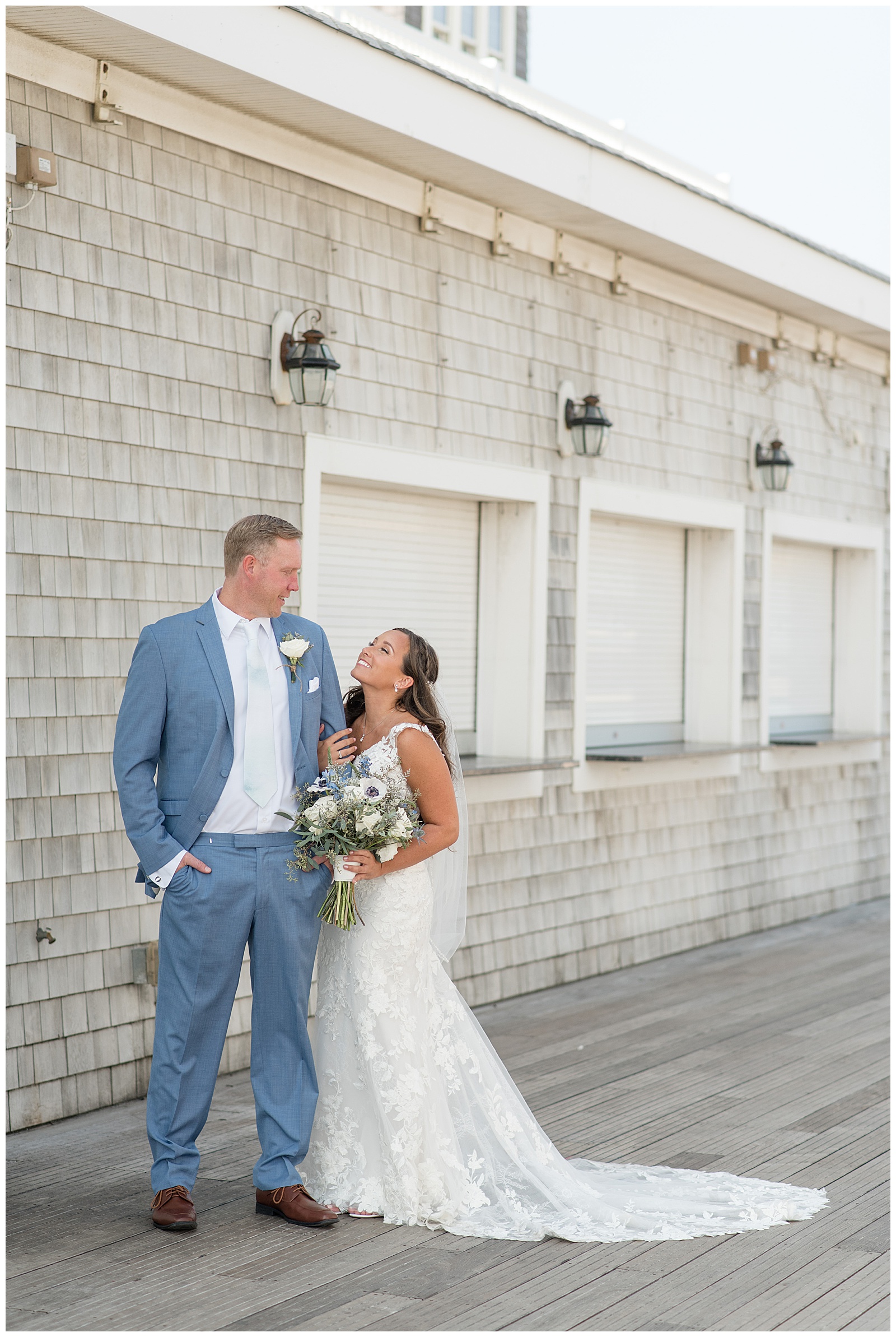 bride and groom standing close as bride looks up at her tall groom by gray building in Avon by the Sea