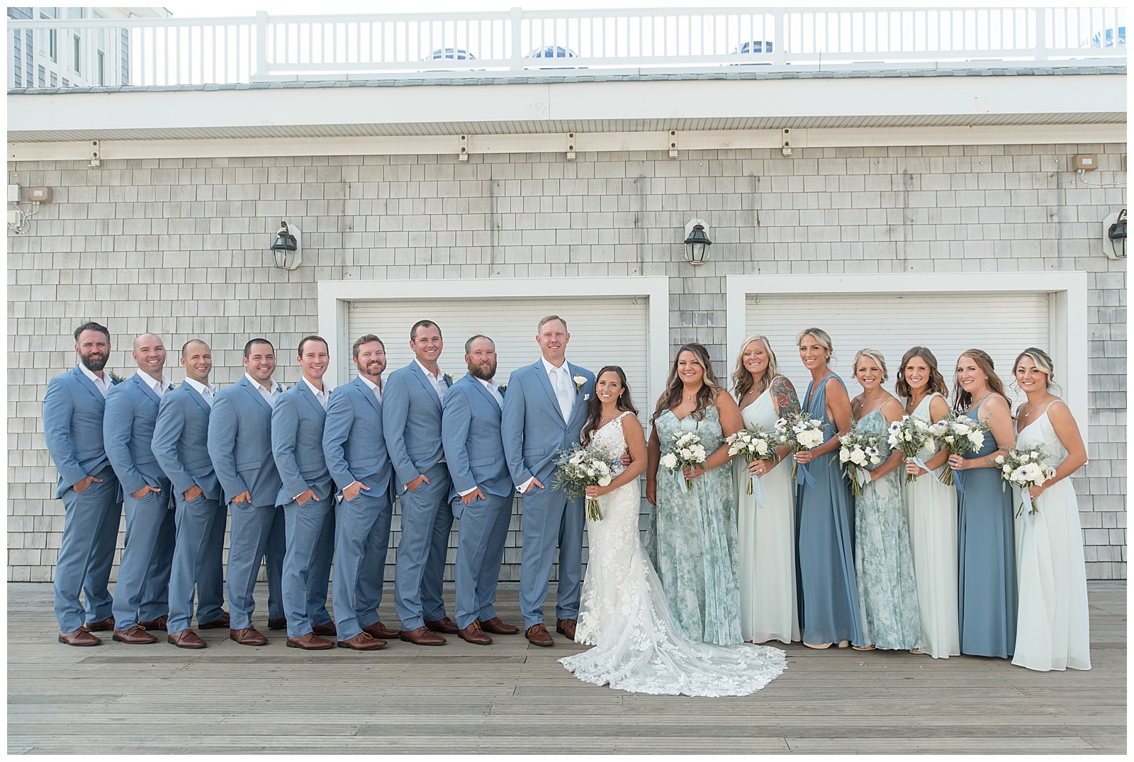 bride and groom surrounded by their large bridal party all wearing shades of blue by gray building in avon by the sea new jersey