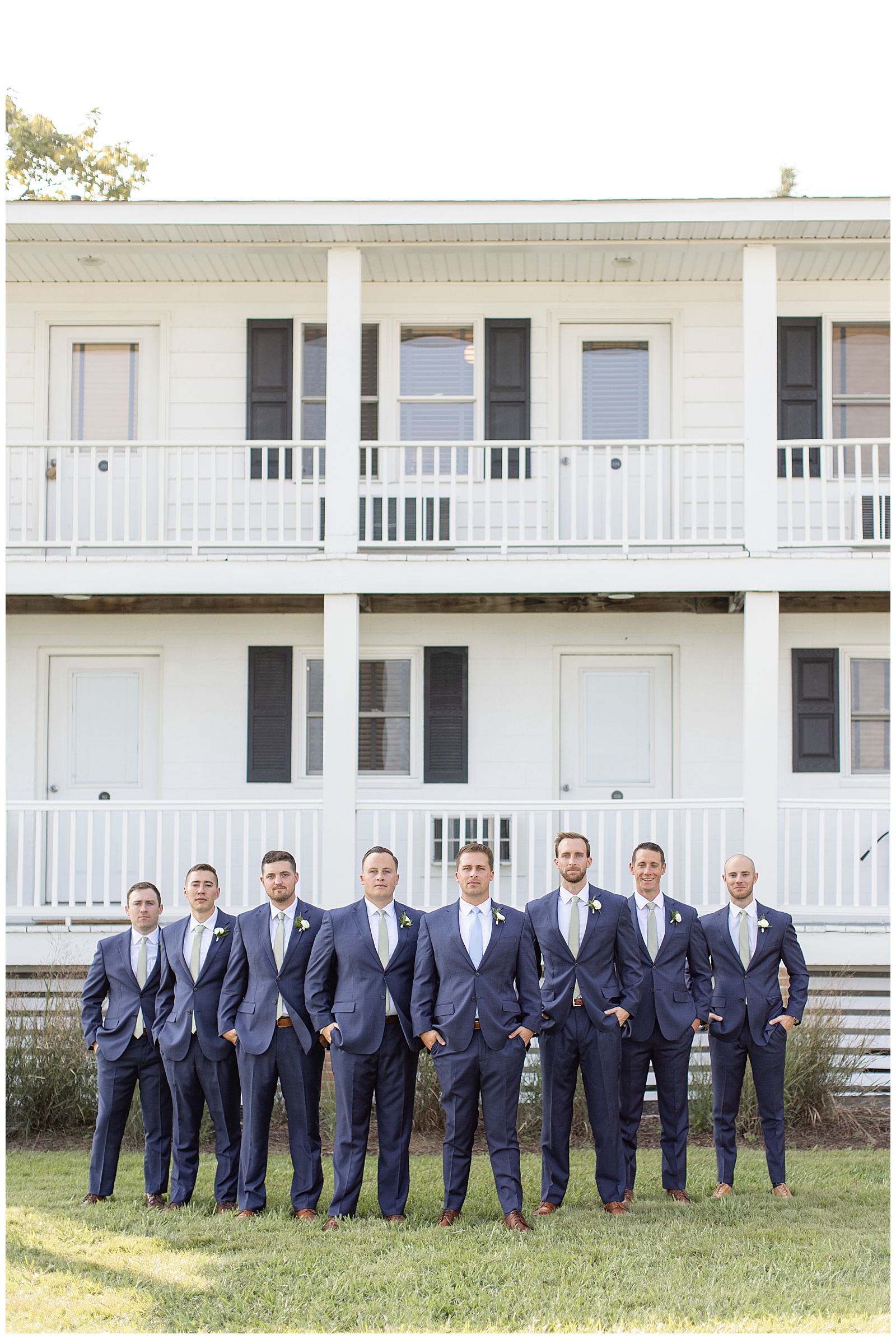 groom with his seven groomsmen all in navy blue suits with hands in pockets by front porch of white beach house in maryland