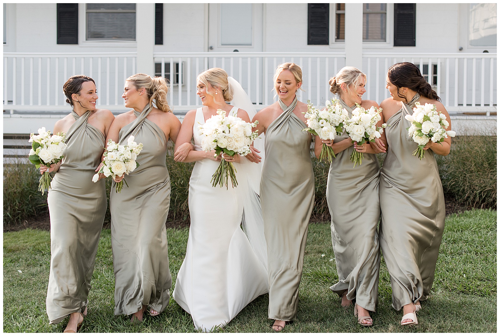 bride and bridesmaids with arms linked holding bouquets and walking towards camera smiling and chatting by front porch on tilghman island