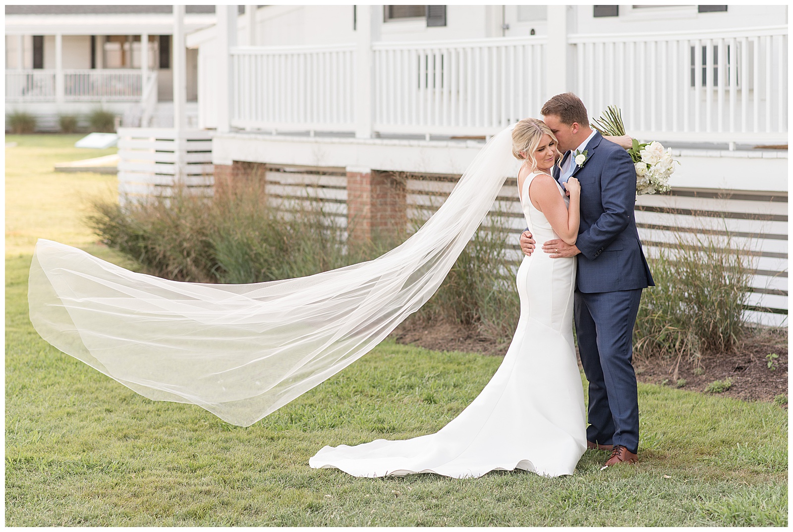 couple on right side of photo hugging as groom nestles into bride's left cheek and long veil blows in breeze in maryland