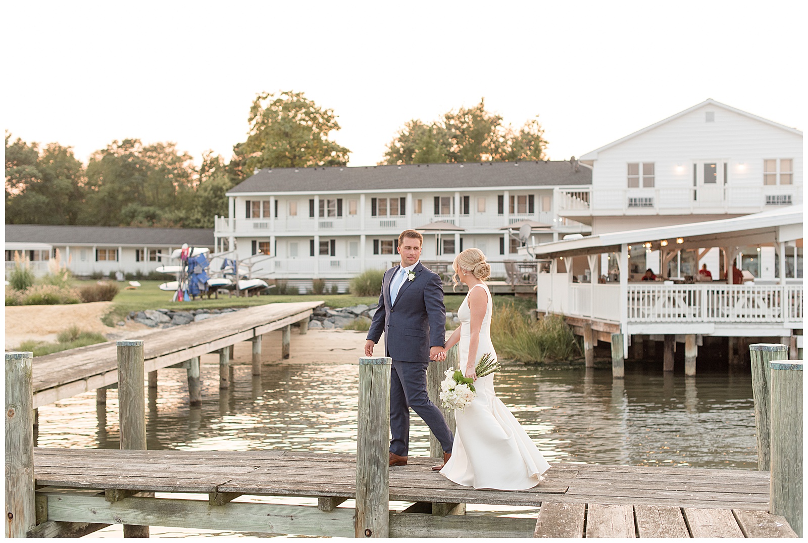 groom holding bride's hand and leading her back along wooden dock with white home in background on tilghman island
