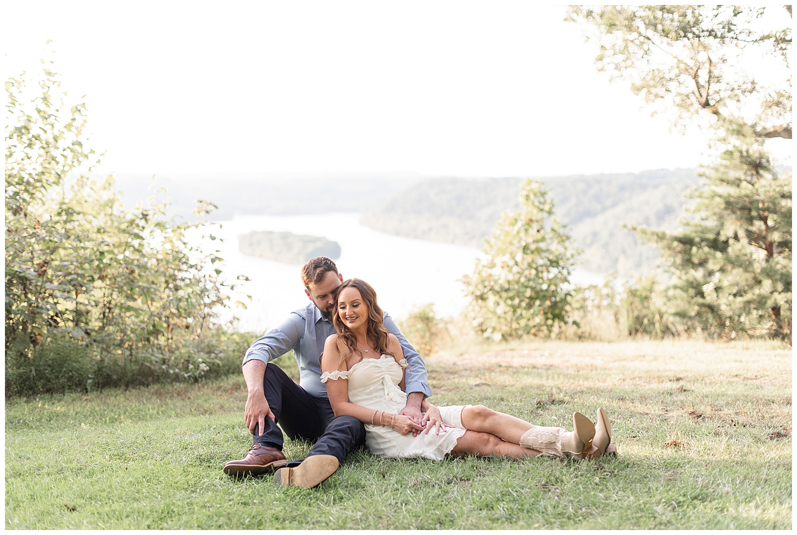 engaged couple sitting in the grass with legs extended and leaning together at pinnacle point by susquehanna river