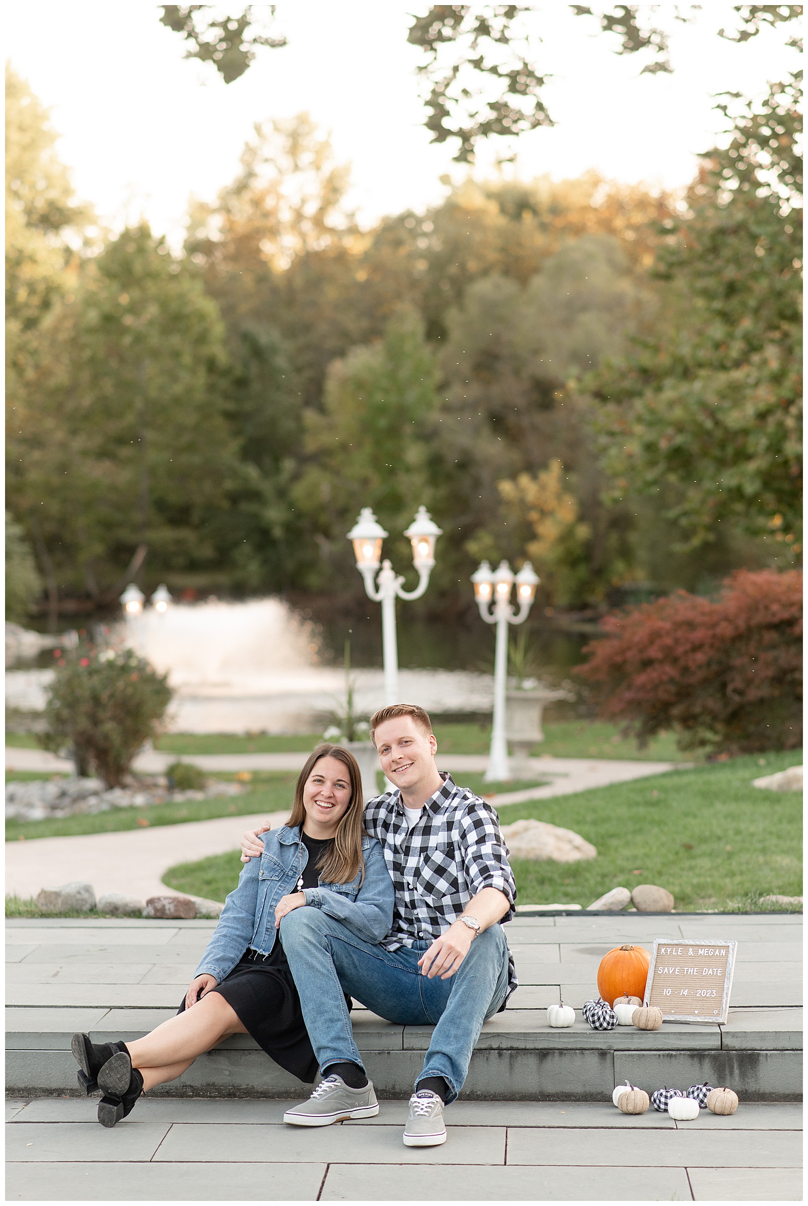 engaged couple sitting on curb with fountain behind them and save the date sign beside them at prophecy creek