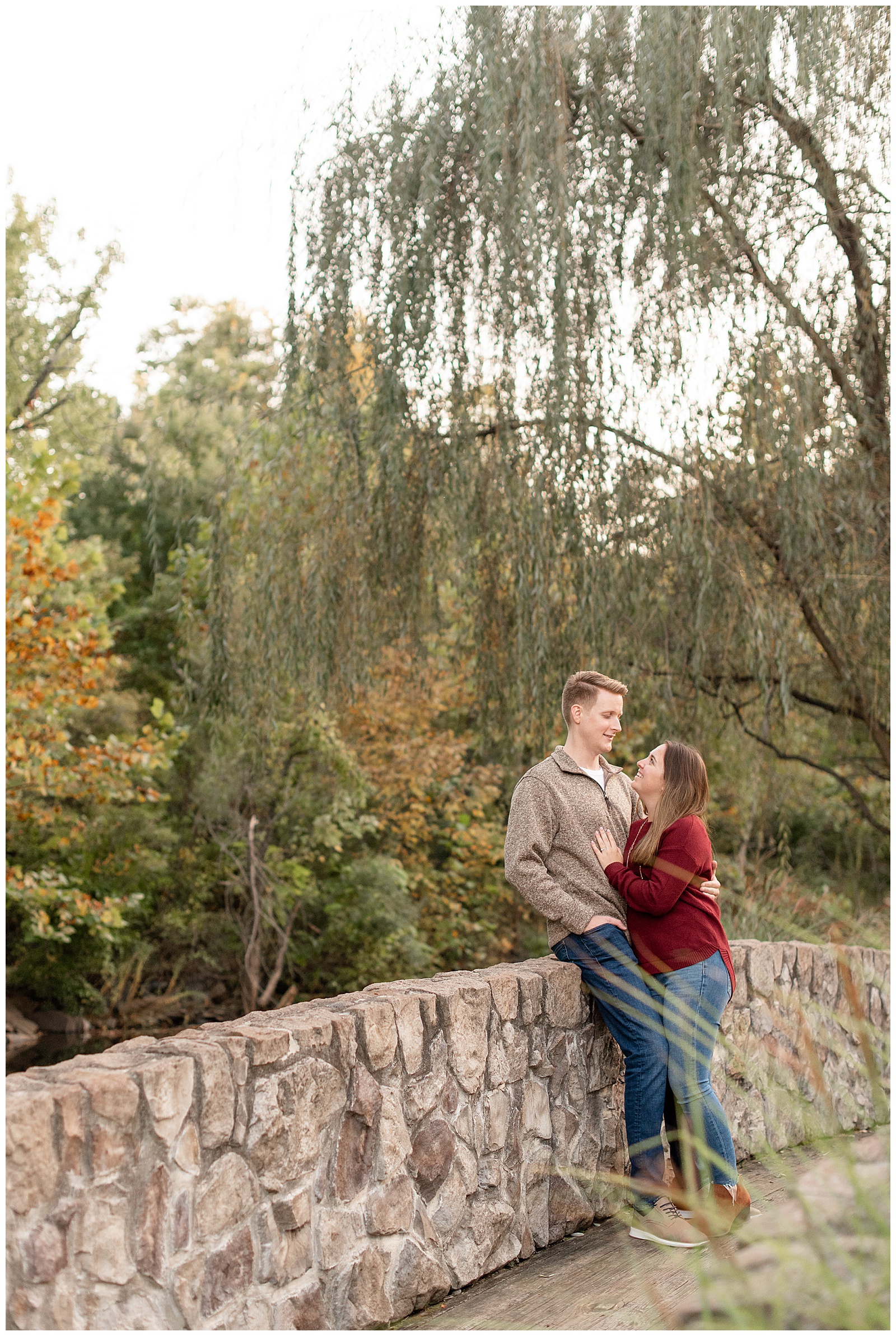 girl wearing maroon sweater and jeans and guy wearing tan pullover and jeans sitting on stone wall by trees in montgomery county