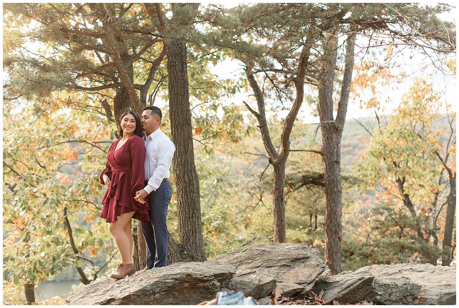 girl in maroon dress and guy in white dress shirt and navy blue pants standing atop huge rock by colorful trees at pinnacle overlook