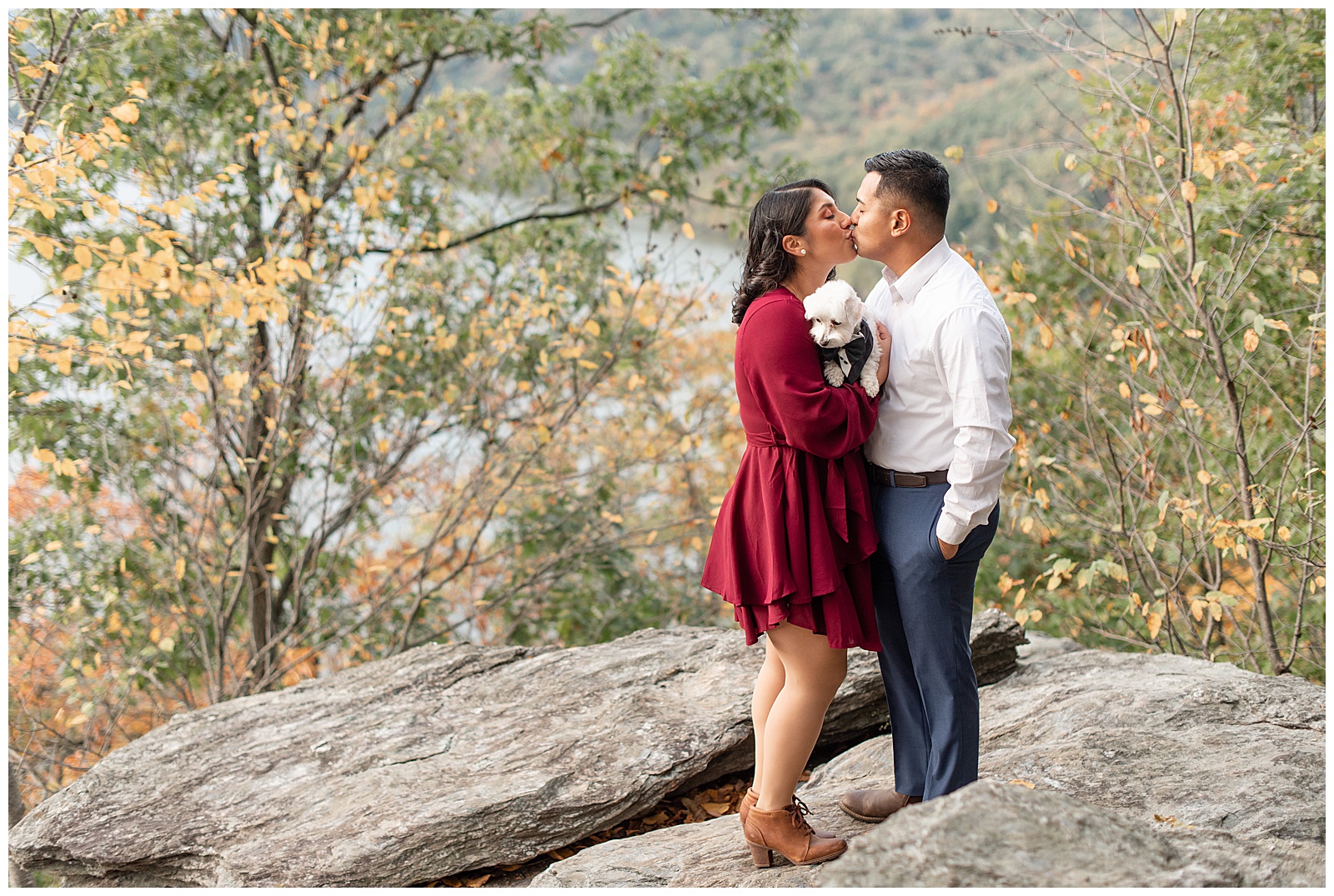 engaged couple kissing atop large rock as they hold their white pup by colorful trees at park in southern lancaster county