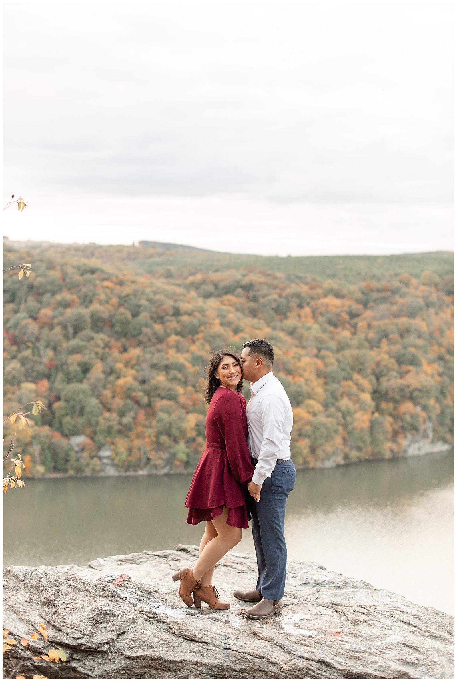 engaged couple holding hands as guy kisses her left cheek at pinnacle overlook with susquehanna river behind them