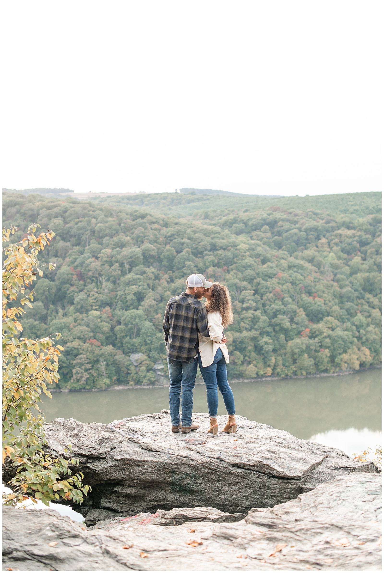 engaged couple standing atop large rock kissing with their backs toward camera by susquehanna river in holtwood pennsylvania
