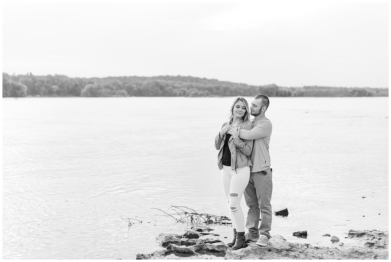 black and white photo of guy standing behind girl and wrapping arms around her along the susquehanna river in lancaster county