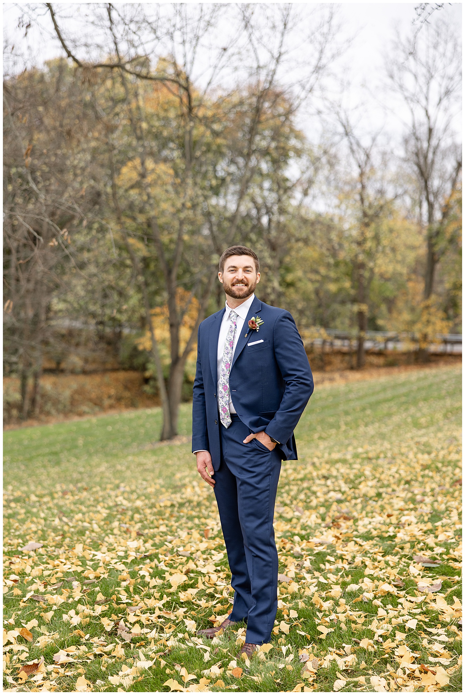 groom wearing navy blue suit standing with left hand in his pocket by colorful fall trees in wrightsville pennsylvania